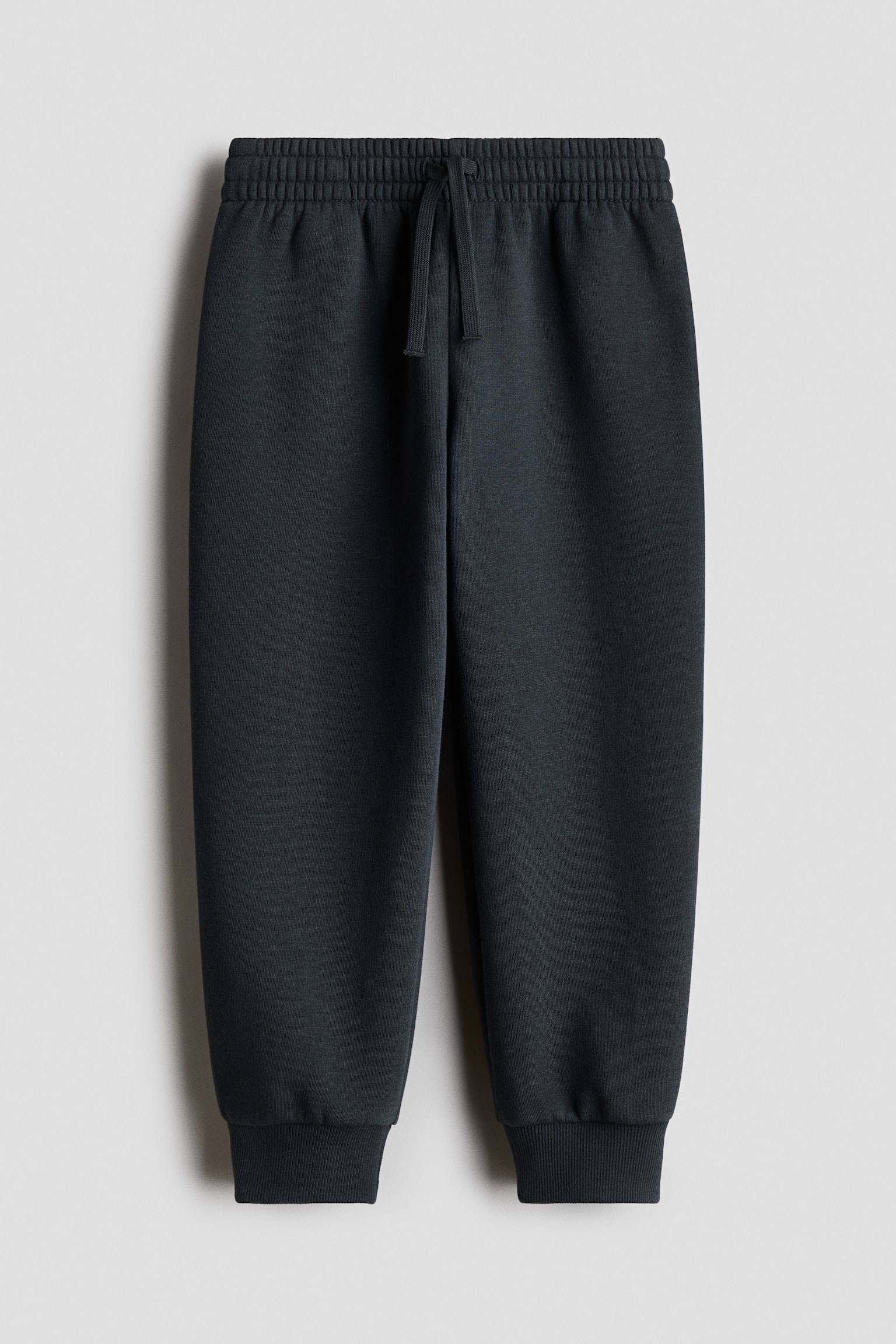 A pair of dark grey track pants is shown from the front, featuring a wide elasticated waistband with a matching drawstring tied at the centre. The pants taper slightly down the legs, ending in ribbed cuffs at the ankles.
