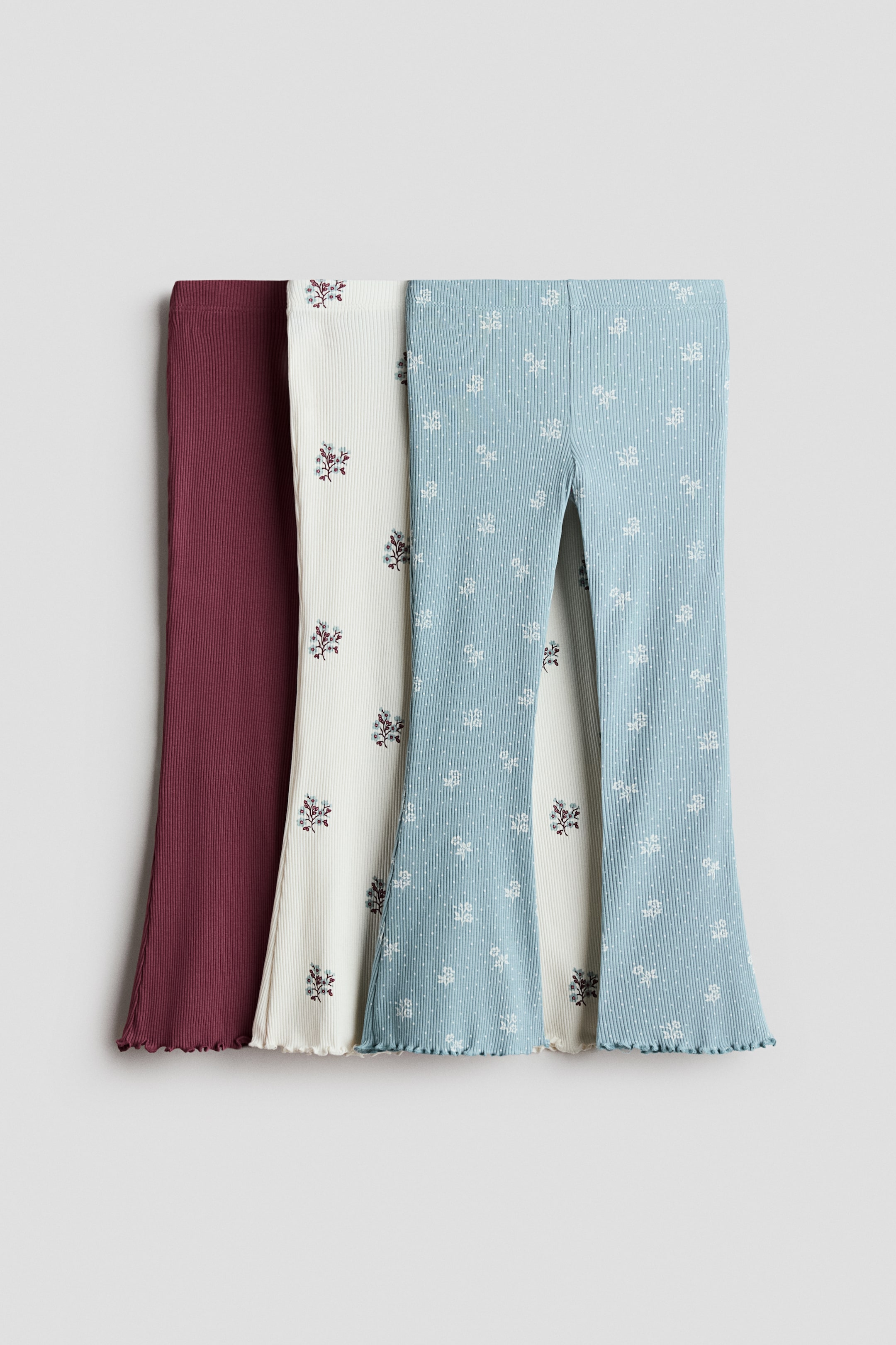 View larger image: Three pairs of ribbed flared leggings are displayed, showcasing their front. The left pair is a solid burgundy, the middle pair is off-white with scattered multi-coloured floral patterns, and the right pair is light blue with white polka dots and scattered white floral patterns. All pairs feature lettuce-edge hems.