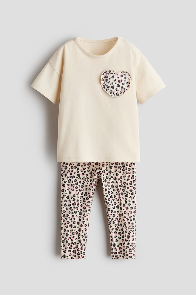 Um conjunto de 2 peças em algodão é apresentado de frente, composto por uma t-shirt de manga curta em tom creme e leggings caneladas a combinar. A t-shirt exibe um bolso no peito com folho em forma de coração, com estampado de leopardo em preto e rosa, enquanto as leggings apresentam o mesmo estampado animal por toda a peça. - 1