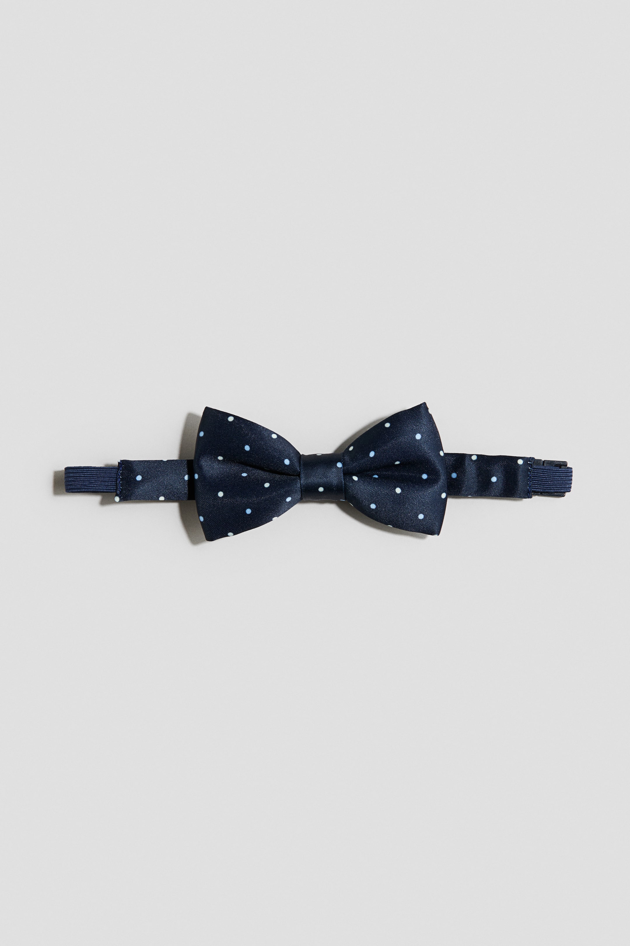 View larger image: "A pre-tied bow tie in dark blue woven fabric with white polka dots is shown front-facing. An adjustable elastic strap extends from both sides, with a plastic fastener visible on the right."