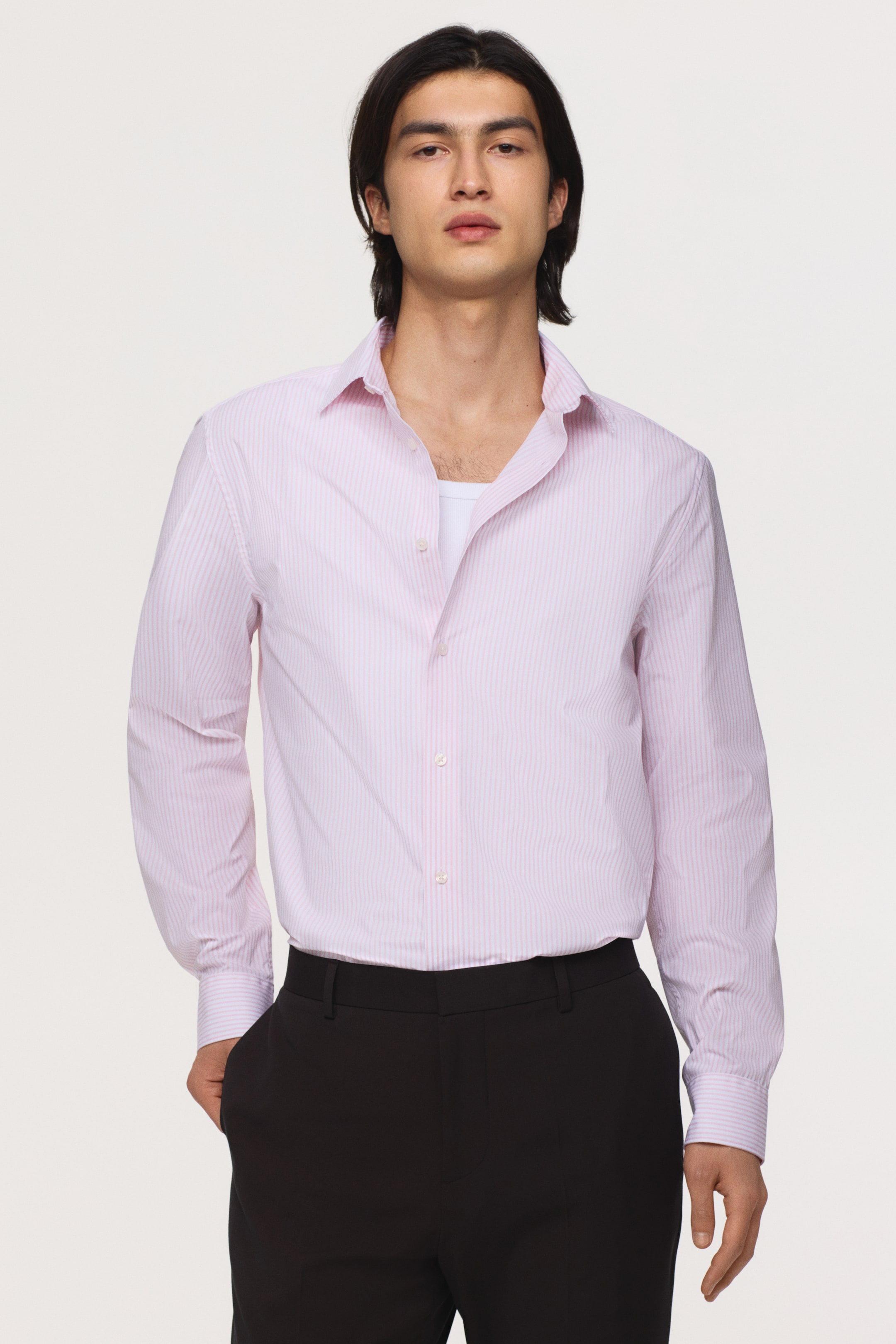 View larger image: The model is wearing a light pink pinstripe shirt featuring a turn-down collar, long sleeves, and buttoned cuffs. The slim-fit shirt is partially unbuttoned to reveal a white undershirt and is tucked into black trousers.