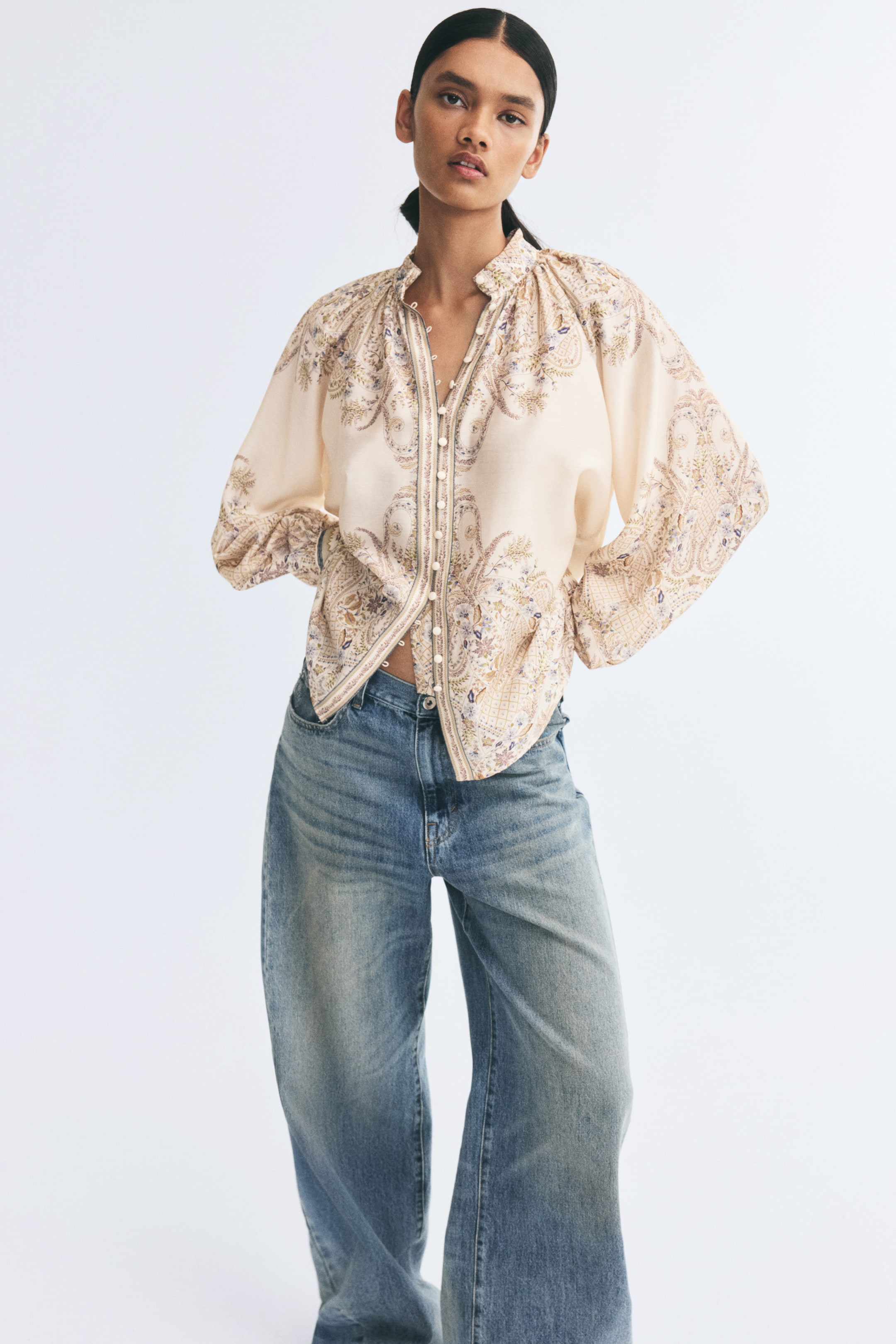 View larger image: A relaxed-fit, cream-coloured blouse with a soft paisley pattern is worn by the model, paired with wide-leg blue jeans. The blouse features a gathered stand collar, a front button closure with covered buttons, and long, voluminous raglan sleeves.