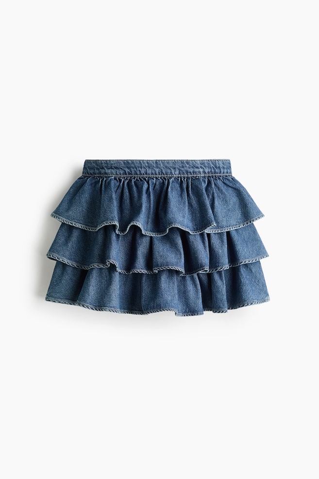 Una falda de mezclilla azul se muestra de frente, con tres niveles de volantes. La falda está diseñada con una pretina fruncida y bordes sutilmente texturizados a lo largo de cada volante. - 1
