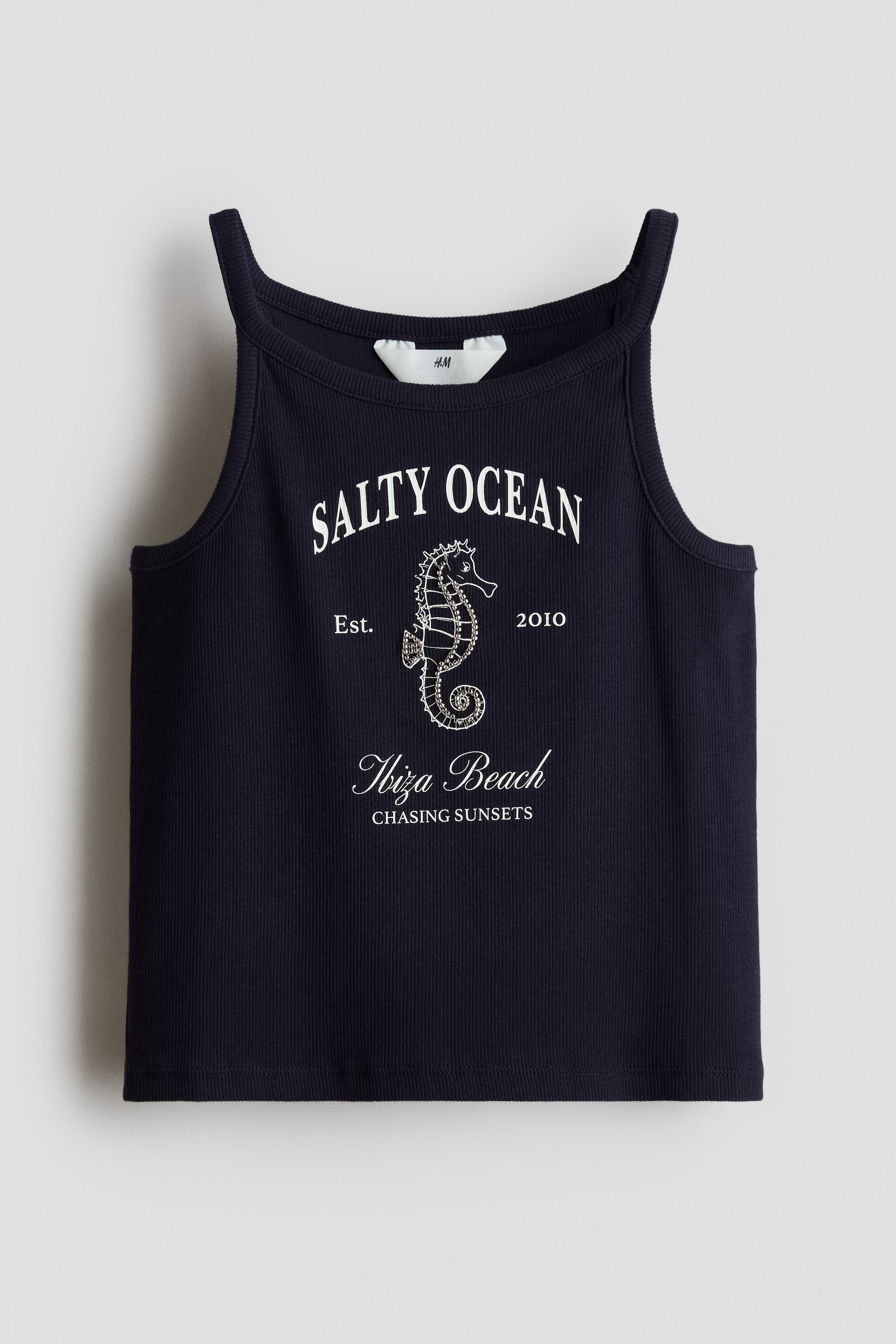 View larger image: A dark blue ribbed knit tank top showcases a white graphic print on its front, featuring "SALTY OCEAN," a seahorse, "Est. 2010," and "Ibiza Beach Chasing Sunsets." The tank top has a rounded neckline with thin straps, and a white H&M label is visible at the back of the neckline.