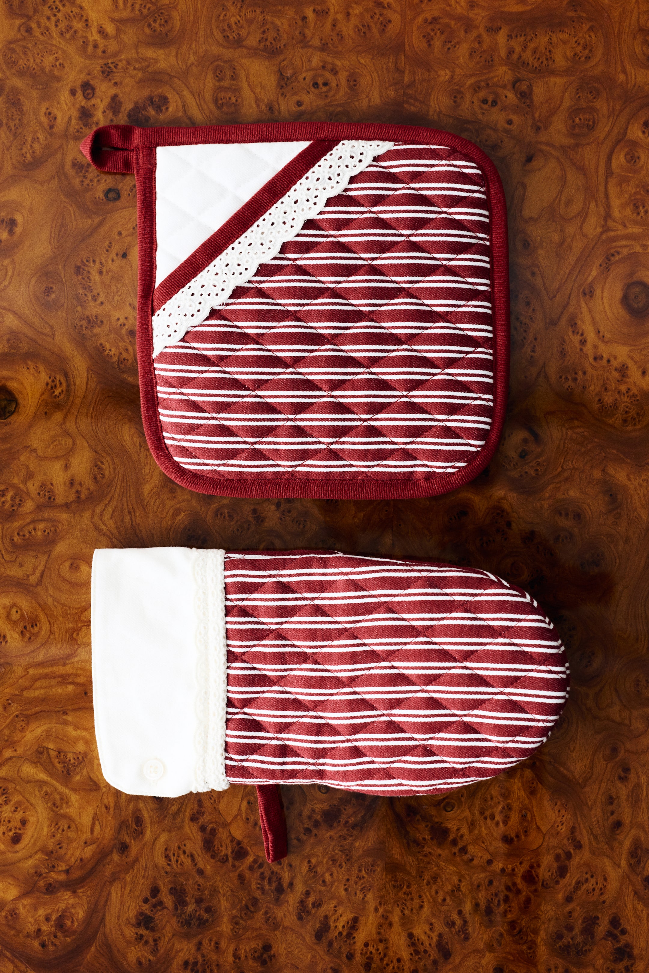 Agrandir l'image: Un ensemble de manique et de dessous-de-plat en sergé de coton matelassé est présenté de face, avec un motif à rayures teintées en fil rouge et blanc agrémenté de délicats détails en dentelle blanche. Le dessous-de-plat carré comporte une poche à main blanche en biais à enfiler et une boucle de suspension, tandis que la manique assortie présente une large manchette blanche unie avec une fermeture par bouton et une boucle de suspension.