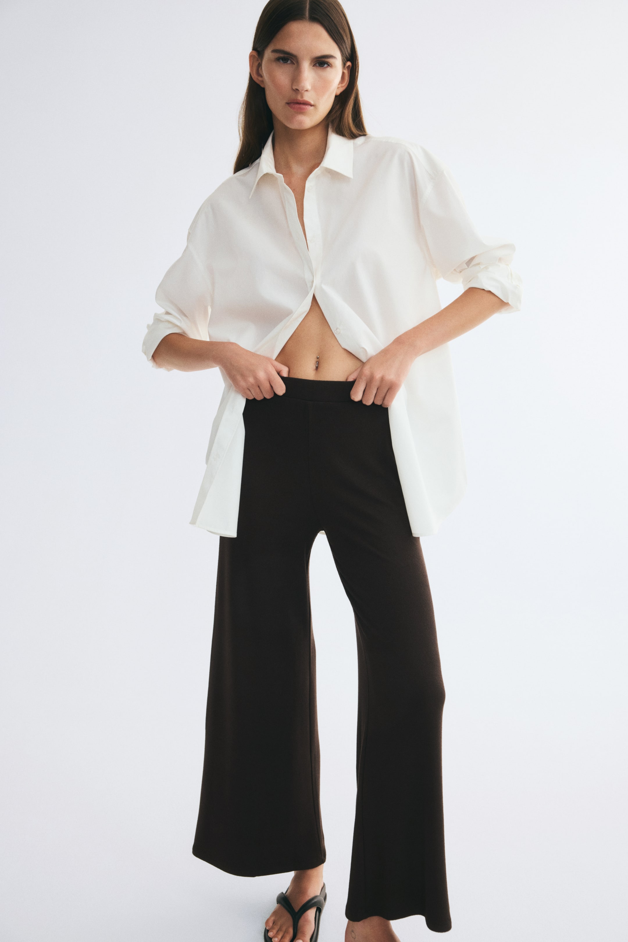 View larger image: Dark brown ribbed jersey pants with a straight-leg silhouette and an elasticated waist are worn by the model, who is holding the waistband with both hands. The loose-fit pants are styled with an open, oversized white button-down shirt.