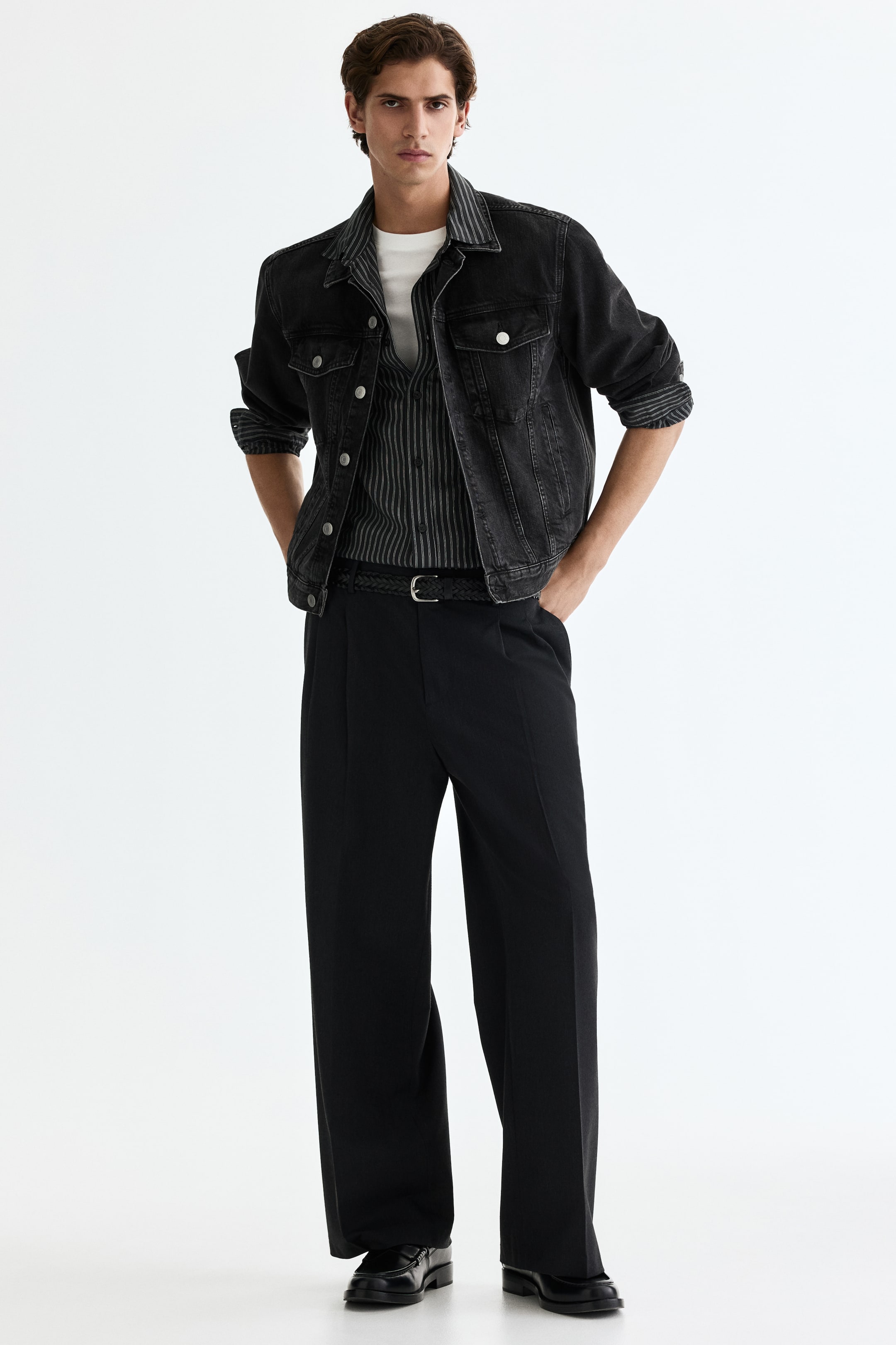 Agrandir l'image: Une tenue est présentée, mettant en vedette une veste en jean délavé foncé avec des poches poitrine et des rabats à boutons, ses manches retroussées pour dévoiler une doublure rayée. La veste est superposée à une chemise à rayures foncées et un t-shirt blanc, tandis que des pantalons noirs à plis larges sont portés avec une ceinture tressée noire et des mocassins noirs.