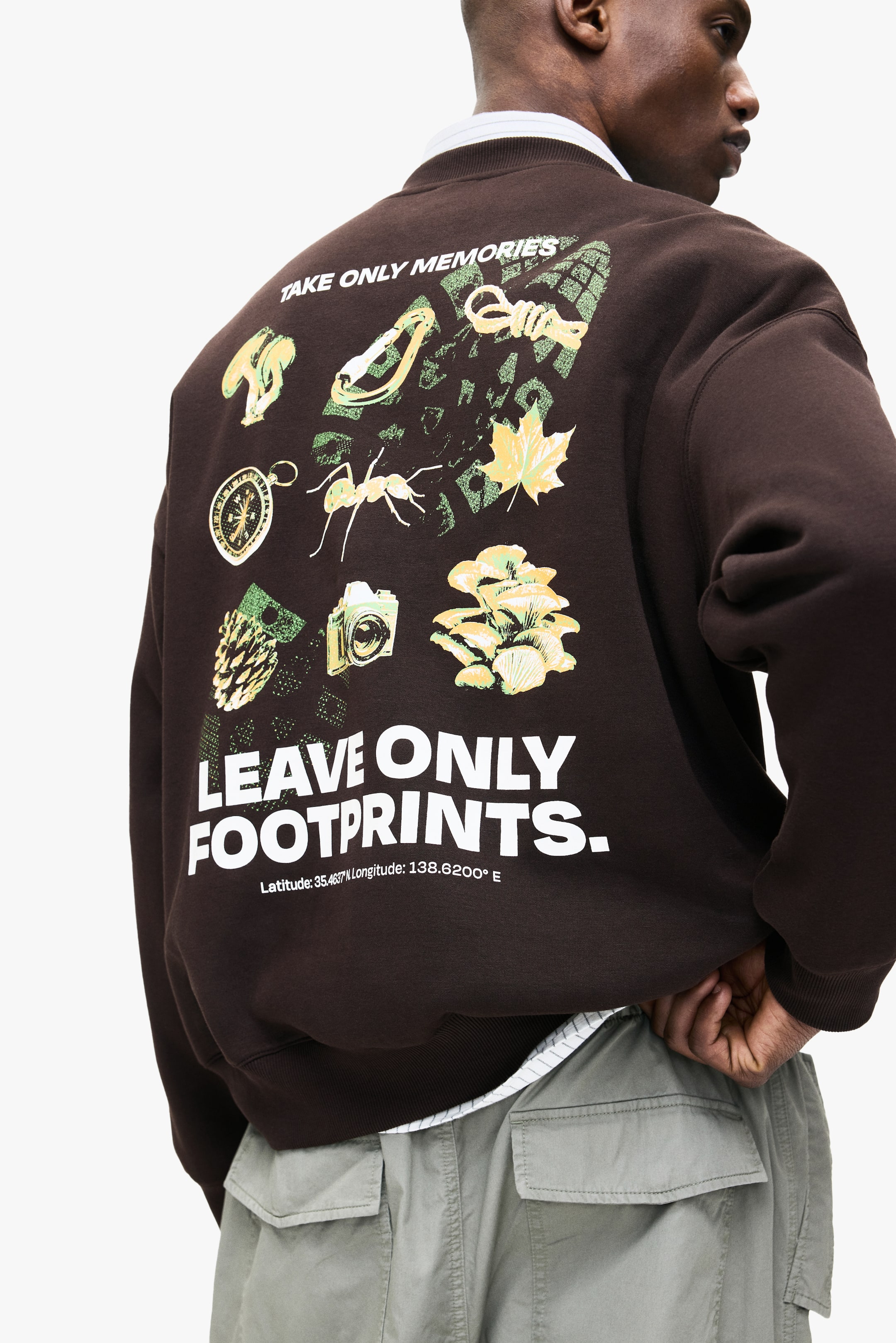 Grösseres Bild ansehen: Ein brauner Crewneck-Pullover, über einem weißen Hemd mit Kragen geschichtet, wird vom Model getragen. Auf der Rückseite des Pullovers sind verschiedene naturbezogene Grafiken und Texte zu sehen, darunter "TAKE ONLY MEMORIES" und "LEAVE ONLY FOOTPRINTS.", während das Model unauffällig den Saum des Kleidungsstücks anpasst.