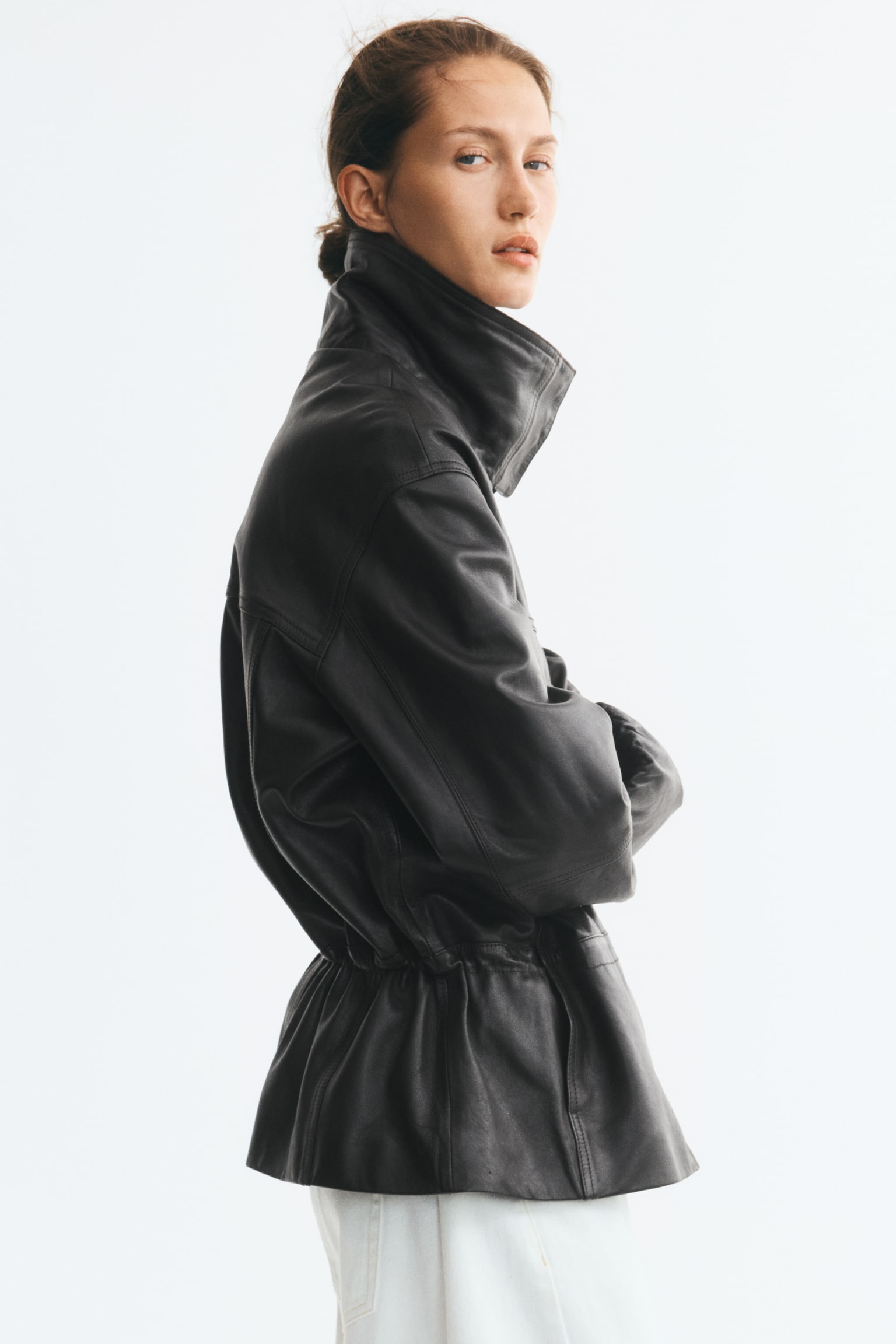 An oversized dark brown leather jacket is shown from the front, featuring a high funnel collar with press-studs and an inner rib-knit collar. It has a two-way front zip, dropped shoulders, elasticated cuffs, flap chest pockets, and a cinched waist with gathered detailing. - 1