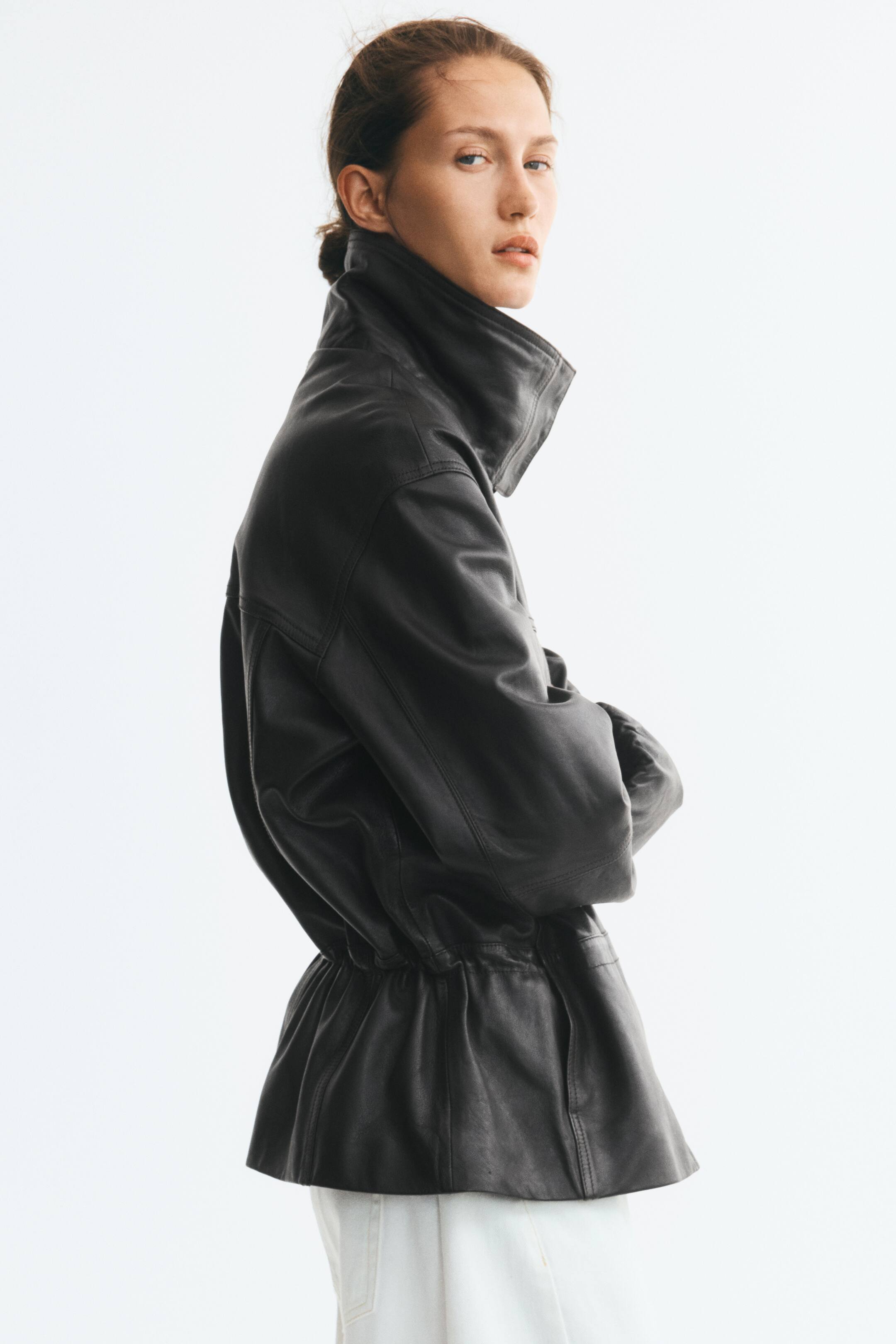 Grotere afbeelding bekijken: Een oversized zwart leren jack met een hoge, gestructureerde kraag en een geplooide, aangesnoerde taille wordt gedragen door het model. Het jack, dat verlaagde schouders heeft, is gestyled met een witte broek terwijl het model in profiel staat met de armen over elkaar.