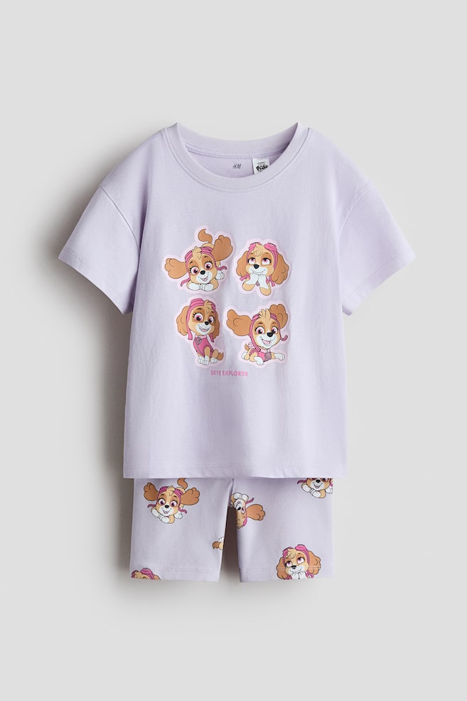 A light purple printed set, consisting of a T-shirt and matching cycling shorts, is displaying on a flat surface. The T-shirt, facing front, is featuring a ribbed trim around the neckline, dropped shoulders, and a graphic print of four Skye cartoon dog characters with 'SKYE EXPLORER' text, while the cycling shorts are showcasing an all-over print of the same Skye characters. - 1