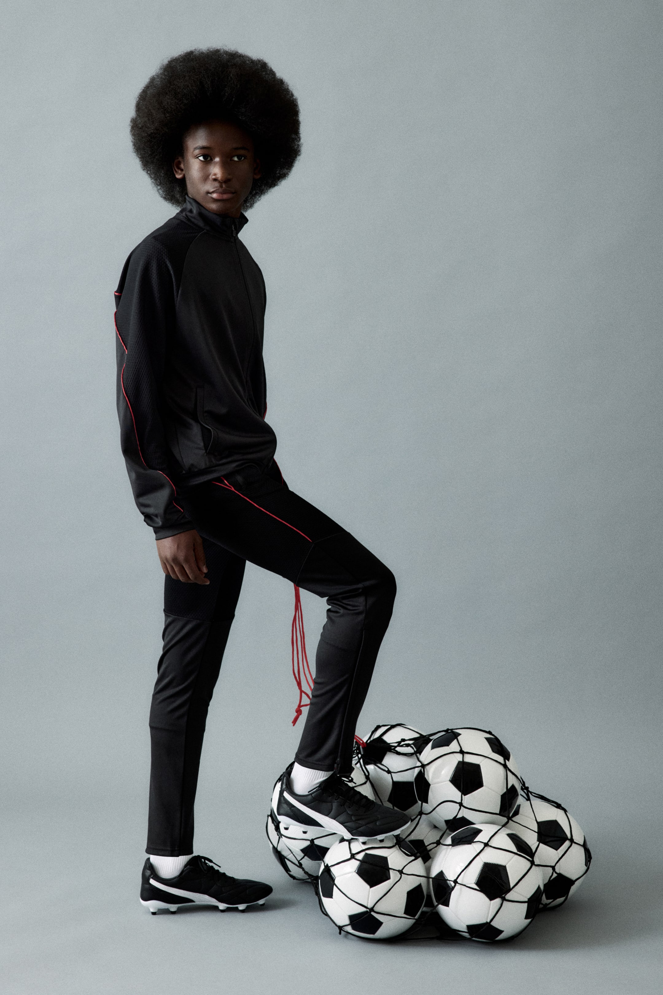 View larger image: A black athletic tracksuit features a jacket with a stand-up collar, a full-zip closure, and red piping along the raglan sleeves and sides. The matching slim-fit track pants feature red piping along the outer seams, mesh-textured panels, and red drawstrings at one hem, with black soccer cleats completing the athletic ensemble.