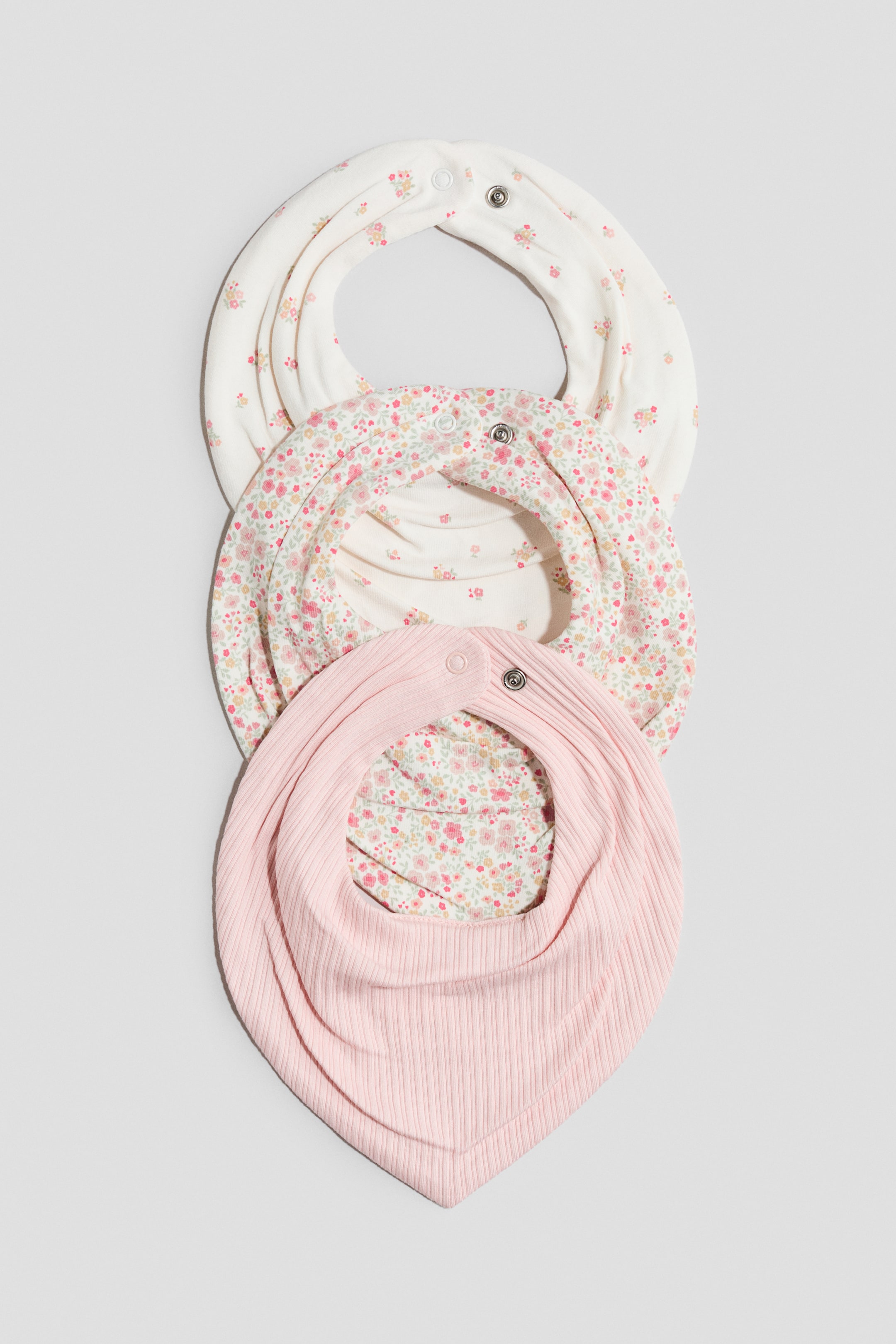 Ver imagen más grande: Tres baberos triangulares de doble capa, hechos de un jersey acanalado y supersuave, se muestran de frente, cada uno con un cierre ajustable de broche a presión en la parte posterior del cuello. Los dos baberos superiores exhiben tela color crema con pequeños patrones florales, mientras que el babero inferior presenta un exterior rosa sólido con un forro floral.
