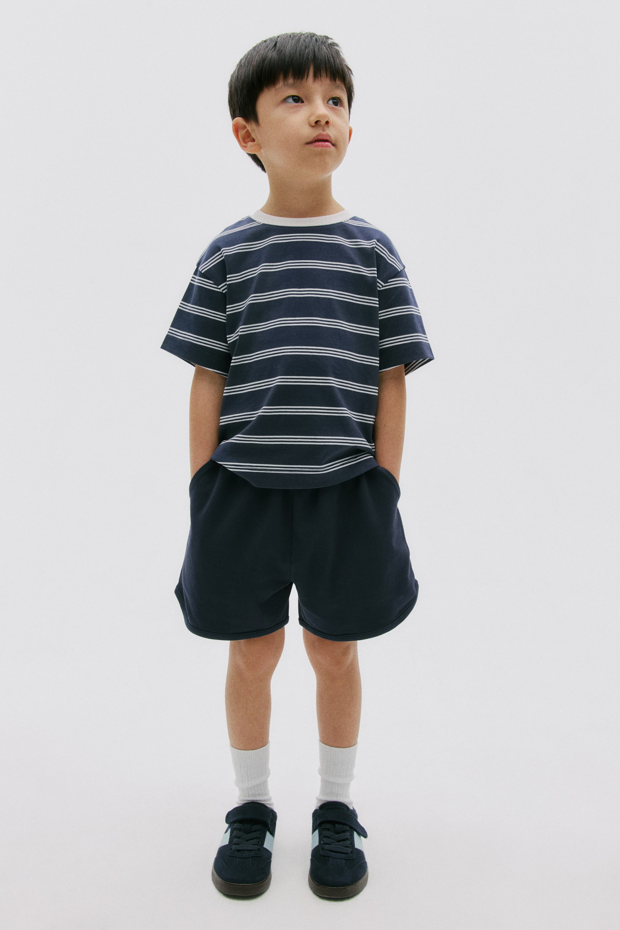 拡大画像を表示: ネイビーブルーのクルーネックTシャツには、細い白の横縞があります。ネイビーブルーのショートパンツも履いており、モデルの手はショートパンツのポケットの中に入っています。