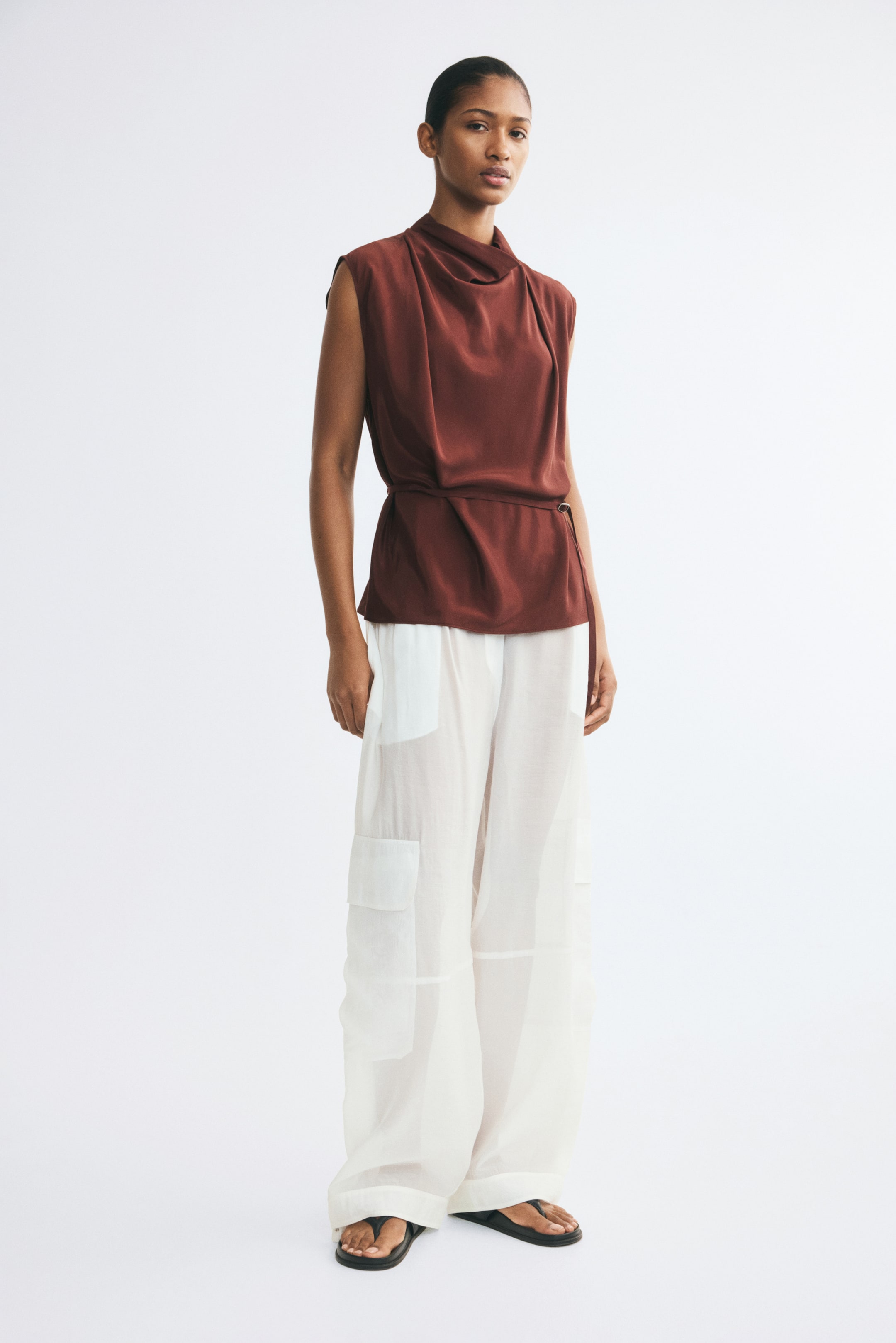 View larger image: A sleeveless rust-brown top is worn by the model, featuring a draped cowl neckline, pleated shoulders, and a narrow adjustable waist belt with a metal slide buckle. The top is styled with semi-sheer white wide-leg trousers and black flat sandals.