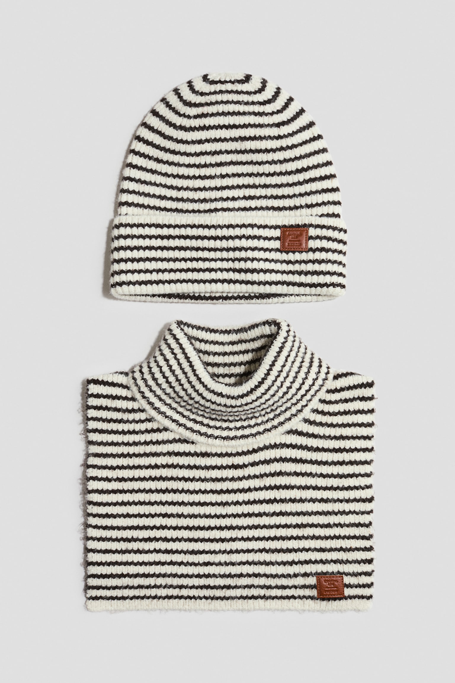 A striped knitted winter accessory set features a matching beanie and neck warmer in off-white and dark brown. The beanie has a folded cuff with a small brown logo patch on the front, and the neck warmer features a high roll-neck collar with a matching patch near the bottom corner.