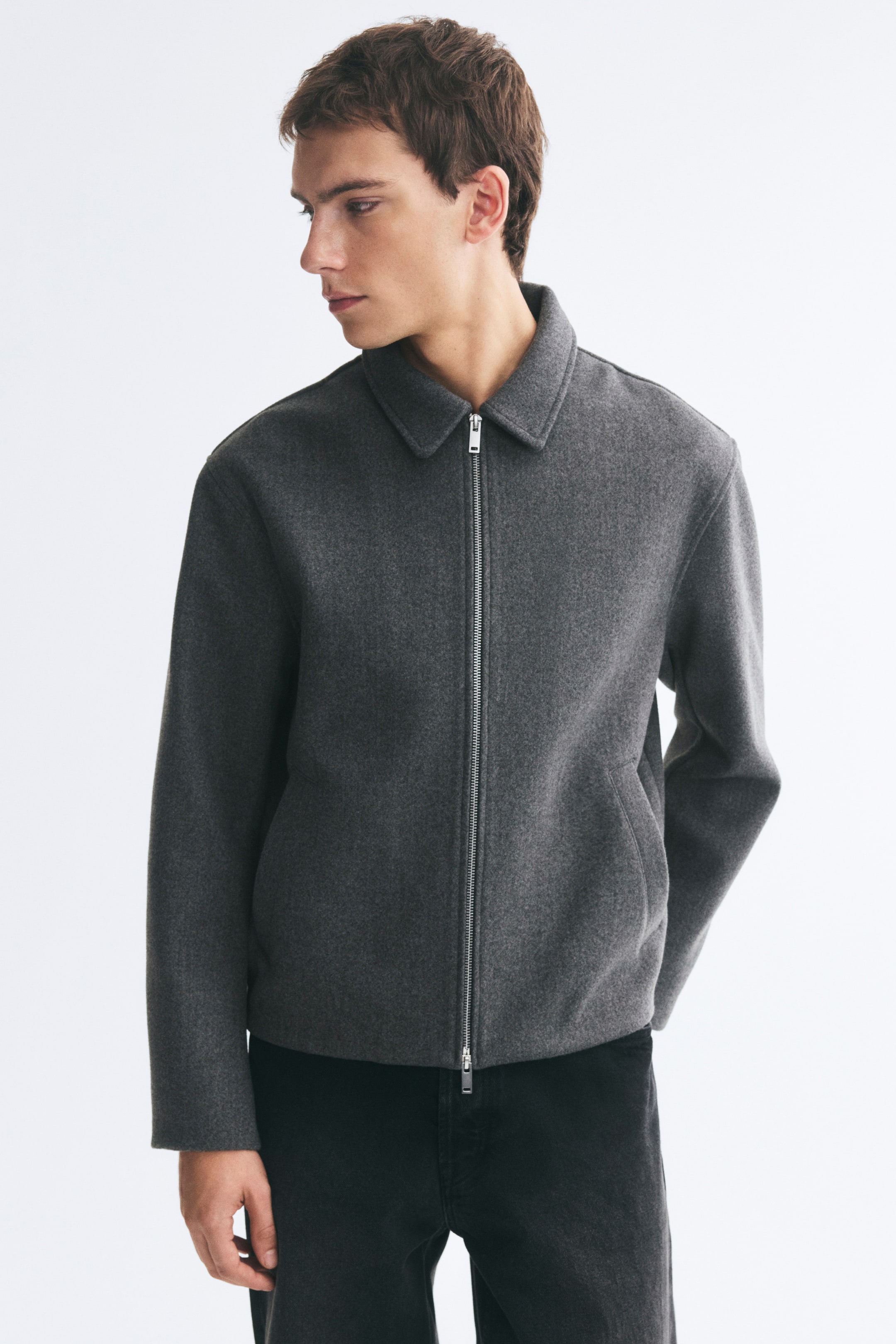 Visa större bild: En overshirt i grått filtad tyg bärs av modellen och har en klassisk krage, en silverfärgad dragkedja framtill samt passpoalfickor i sidan. Overshirten matchas med svarta byxor.