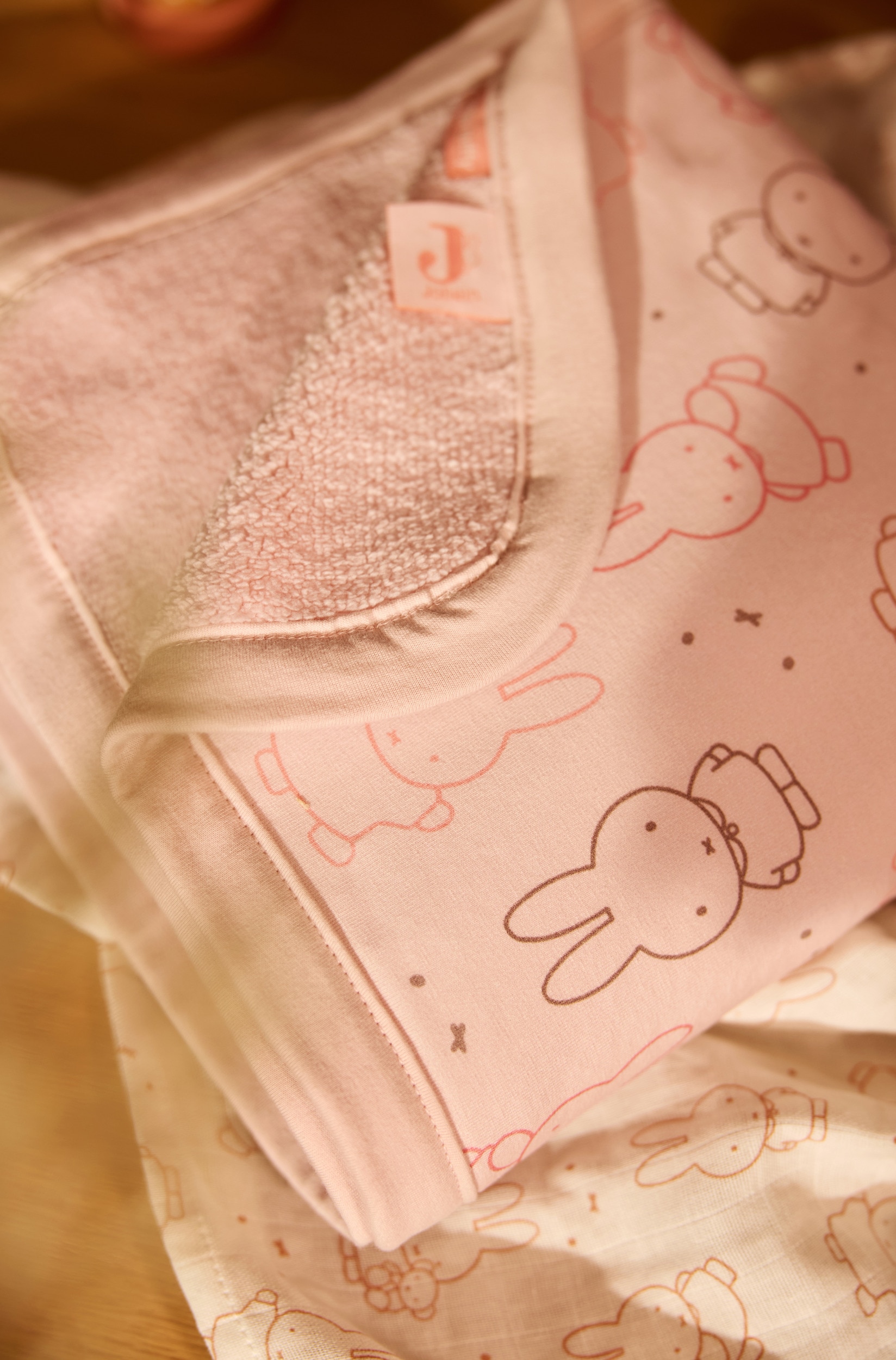 Une couverture pour bÃ©bÃ© rose pÃ¢le pliÃ©e prÃ©sente un motif intÃ©gral de personnages de lapin Miffy dans diffÃ©rentes poses, soulignÃ©s en marron clair et rose foncÃ©. La couverture est dotÃ©e dâun bord uni rose assorti avec finition gansÃ©e, ainsi que dâune petite Ã©tiquette de marque fixÃ©e sur le bord droit.