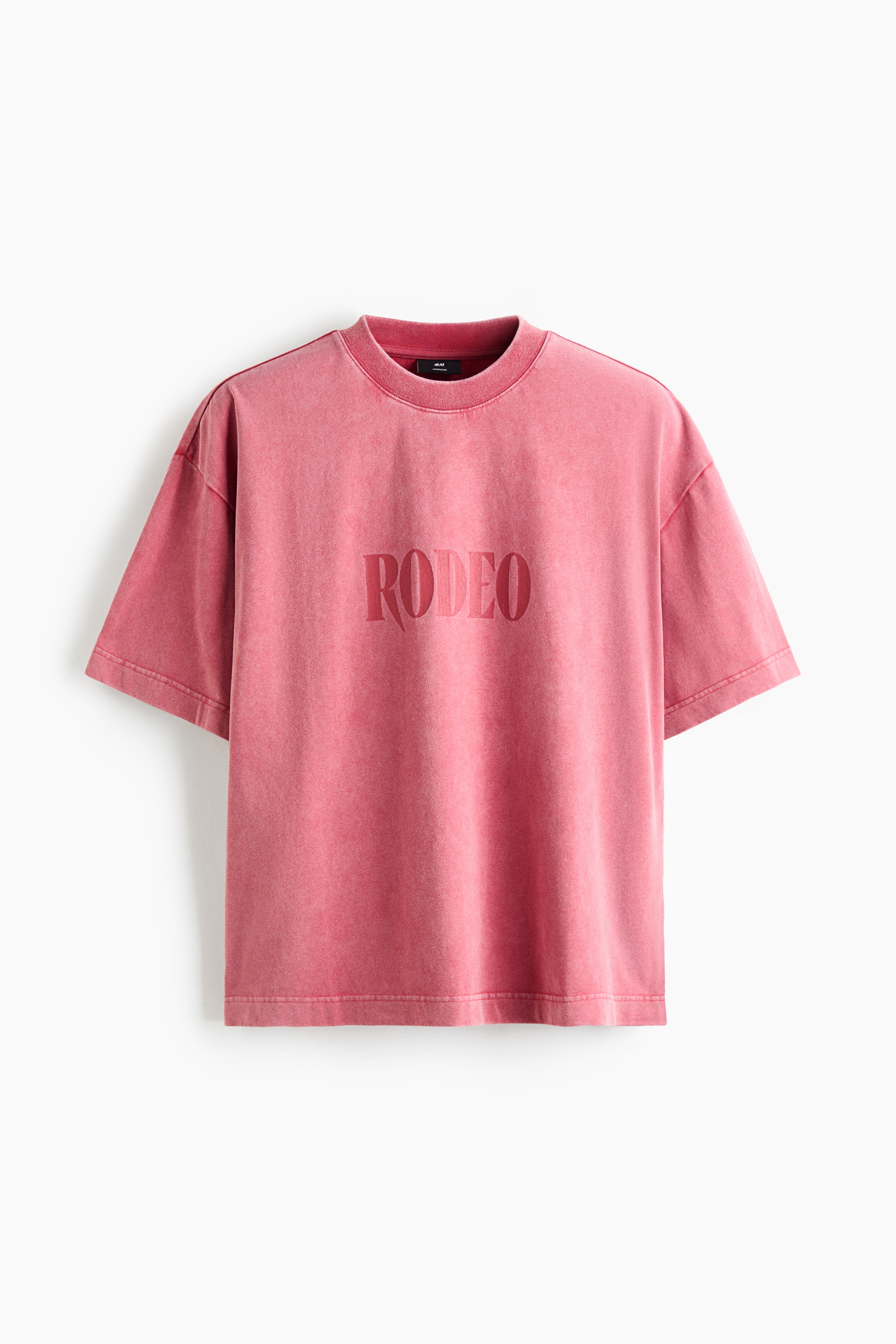 Un t-shirt rose pâle oversize affichant « RODEO » imprimé sur la poitrine est porté par le mannequin. Ce t-shirt en coton épais présente un col rond côtelé, des épaules tombantes et un ourlet droit, et il est porté avec un jean bleu foncé.