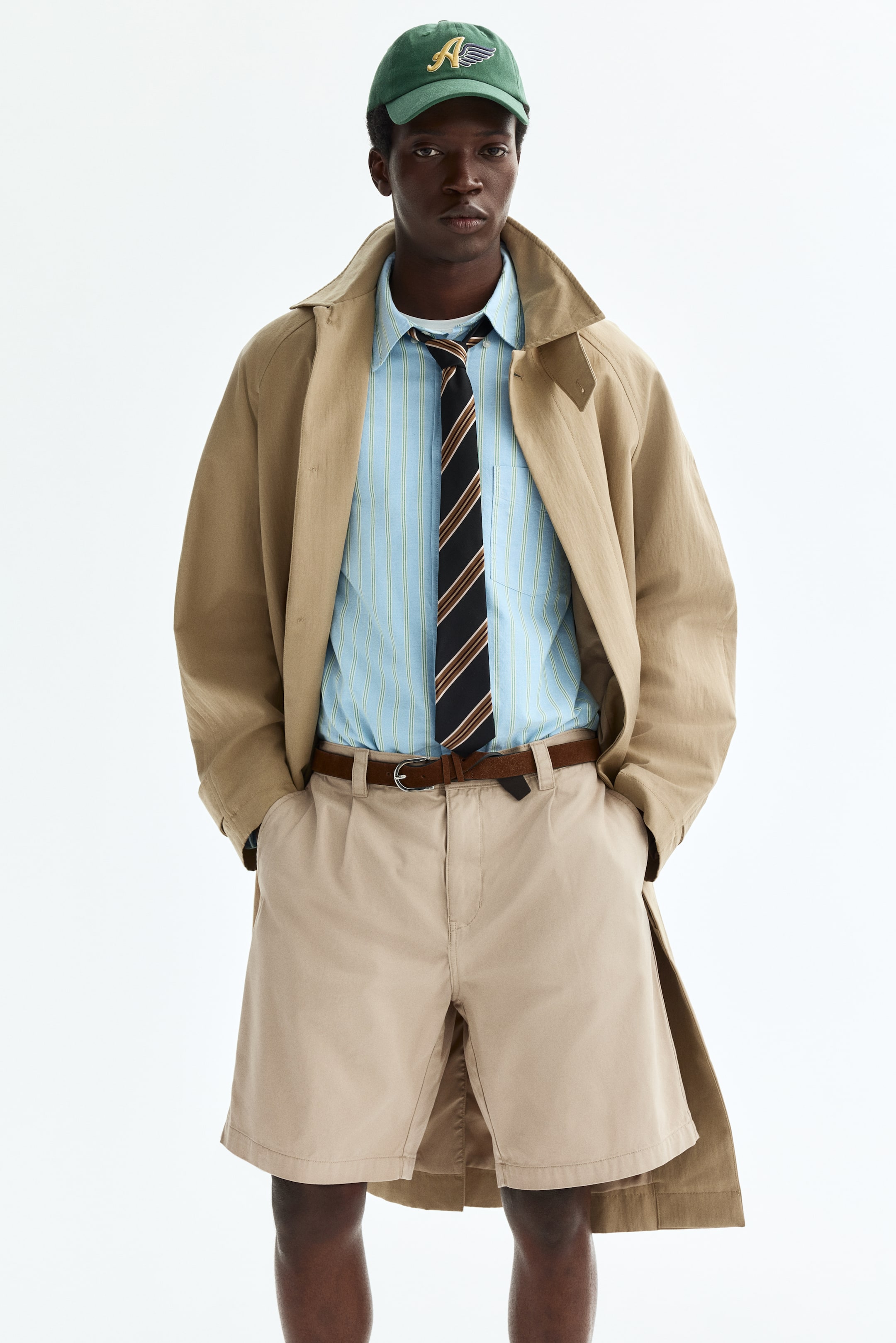 Zobacz większy obraz: Beżowy trencz noszony jest rozpięty na jasnoniebieskiej koszuli z pionowymi paskami, która jest zestawiona z ciemnym krawatem w paski. Beżowe szorty z zakładkami są zabezpieczone brązowym paskiem, a zielona czapka z daszkiem z żółtym logo "A" w kształcie skrzydeł dopełnia stylizację.