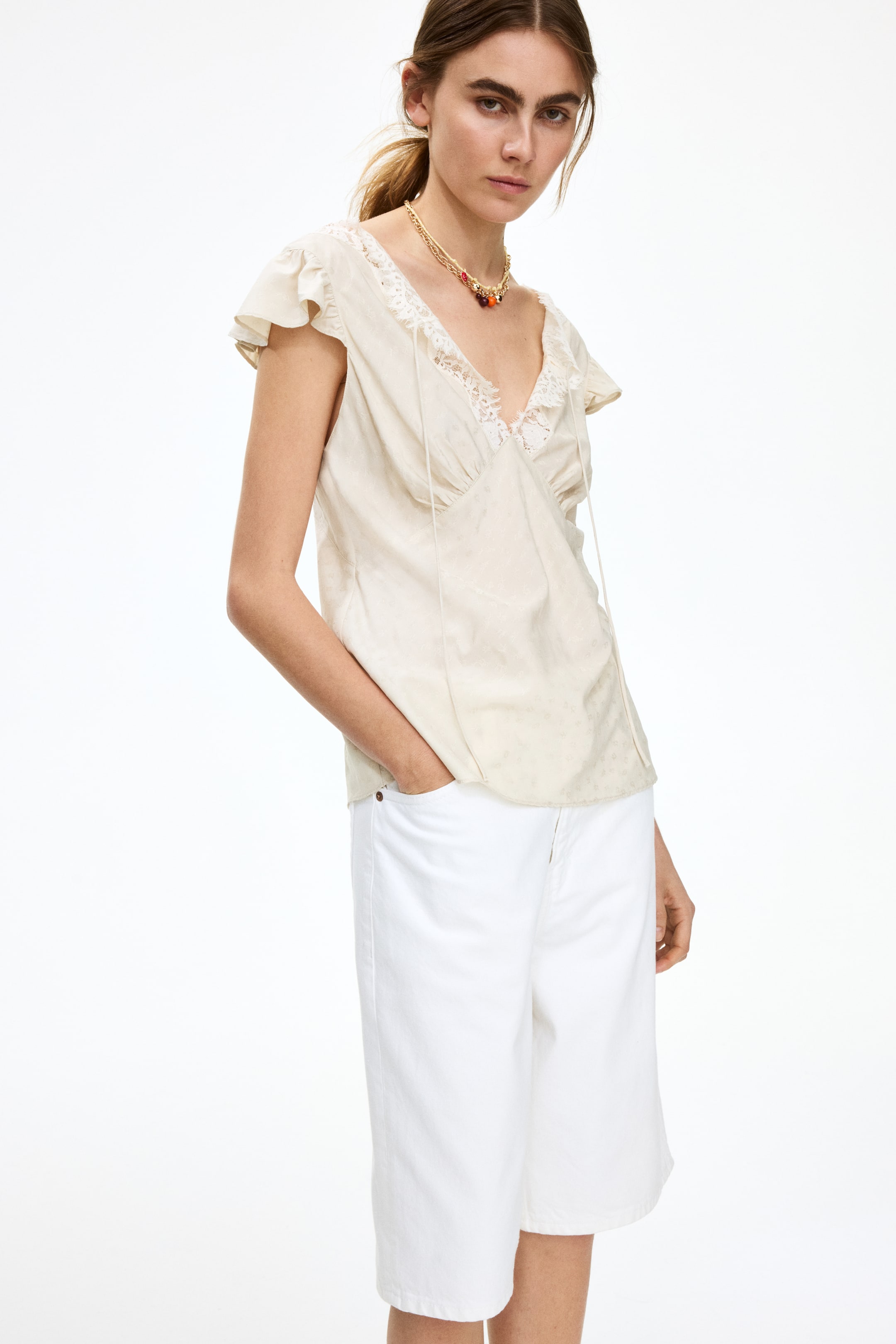 Visualizza immagine più grande: "Una blusa bianco sporco presenta uno scollo a V con bordo in pizzo bianco e un davanti arricciato, da cui pendono cordini da annodare. È progettata con maniche corte arricciate e mostra una delicata fantasia tonale."