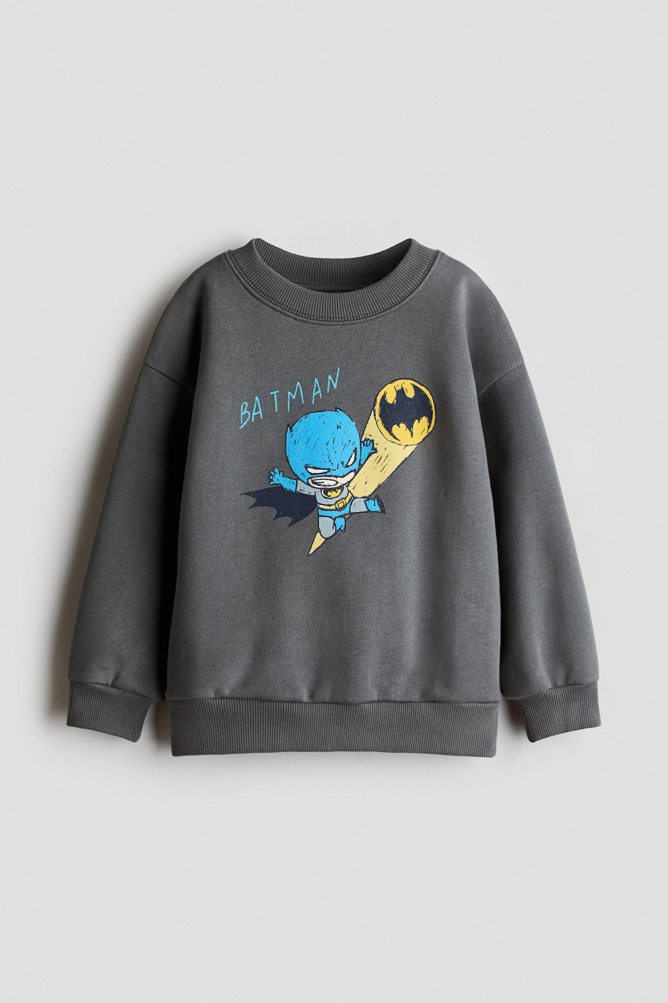 Se større billede: En mørkegrå sweatshirt vises forfra med sænkede skuldre, rund hals med ribkant samt ribbede manchetter og kant. På brystet ses et farverigt trykt motiv af en stiliseret tegneserie-Batman i blå dragt, der flyver langs en gul Bat-Signal-stråle, med ordet "BATMAN" skrevet i blåt.