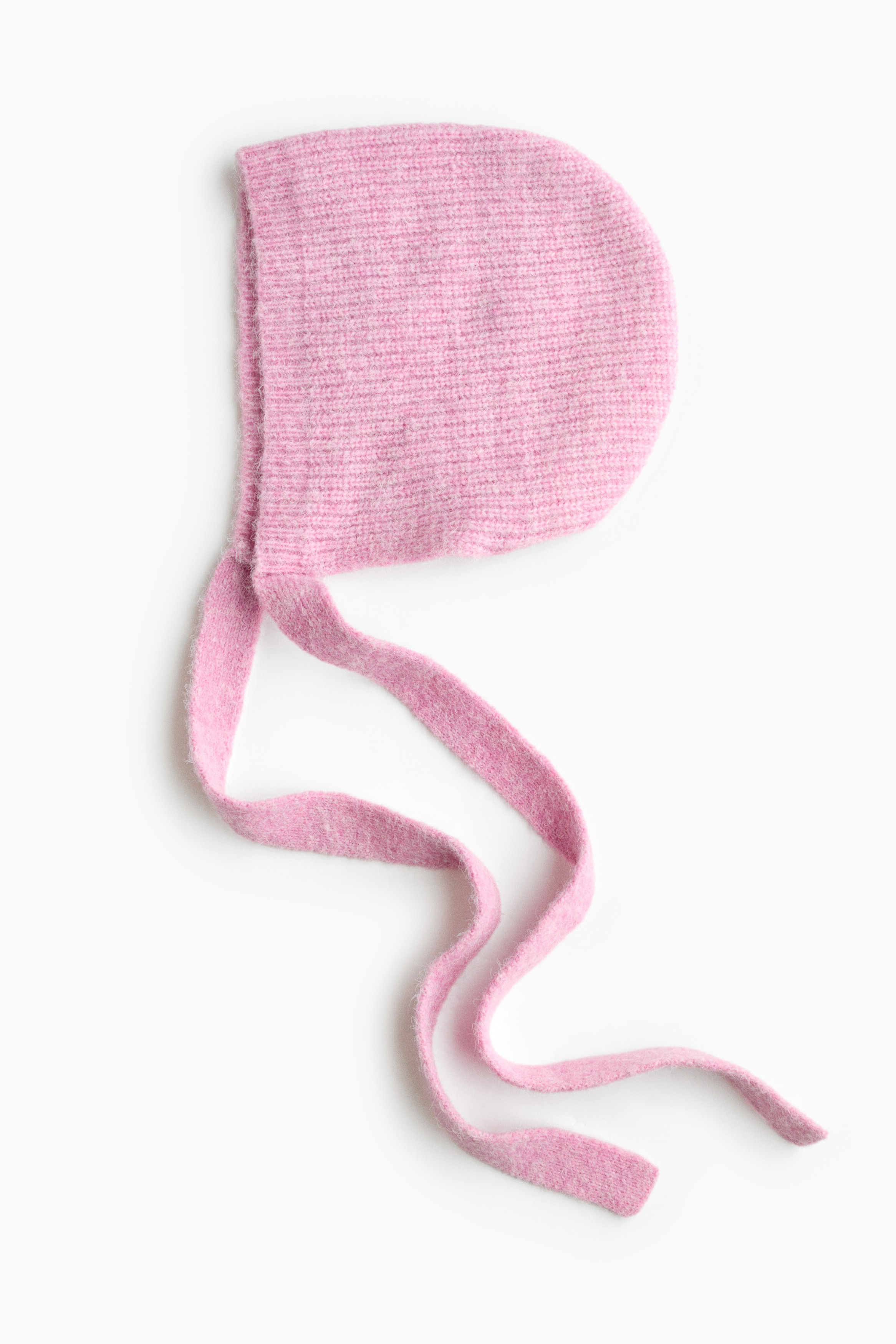 A pink knitted baby bonnet is lying flat in side profile, showcasing a soft ribbed texture and a rounded shape. Two long, matching knitted chin straps extend downward from the bottom front corners in a wavy drape.