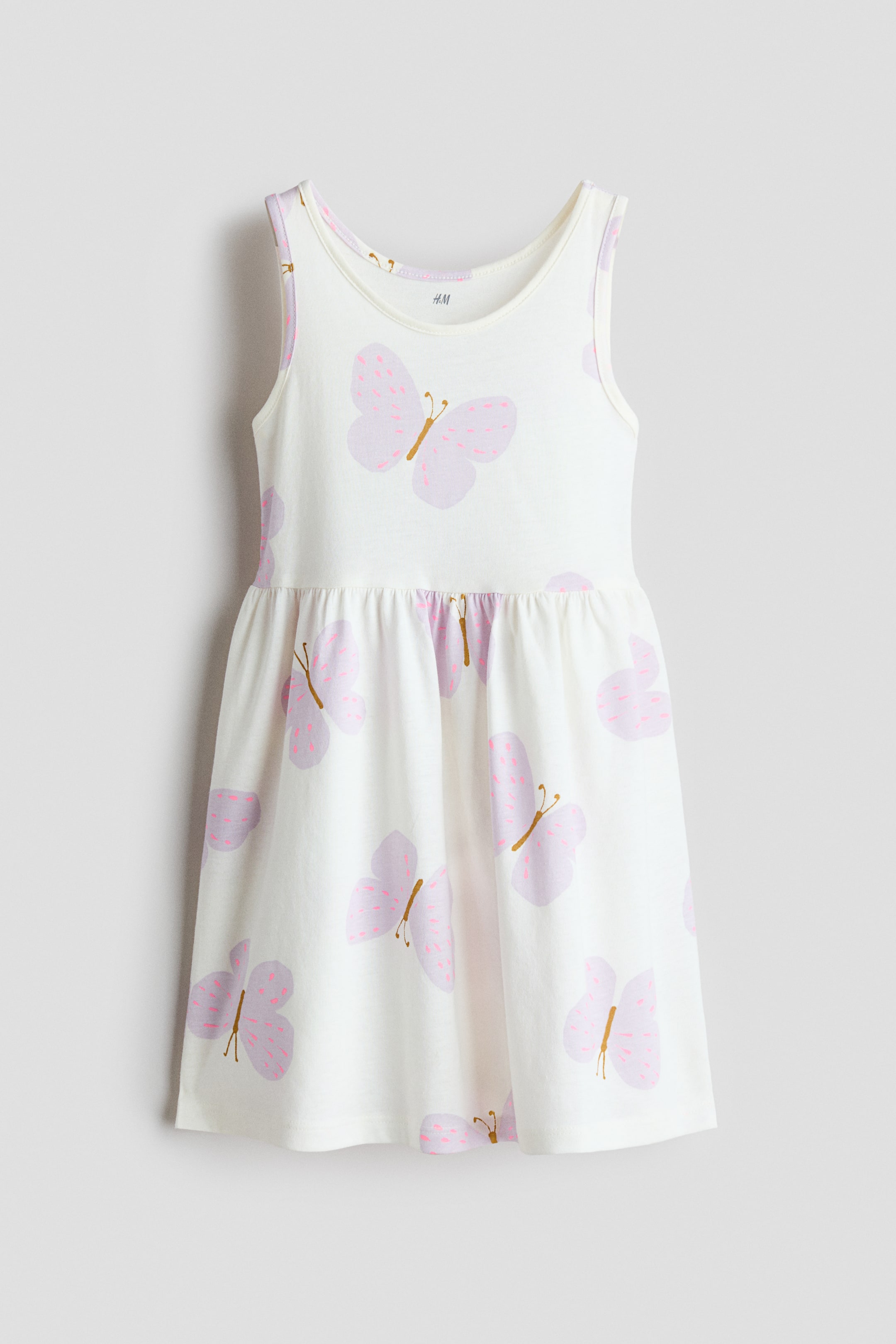 Ver imagem maior: Um vestido branco sem mangas em jersey de algodão exibe um padrão repetido de borboletas lilás claro com pontos rosa e corpos dourados, apresentadas de frente. Apresenta um decote redondo, uma costura franzida na cintura e uma saia suavemente evasê.