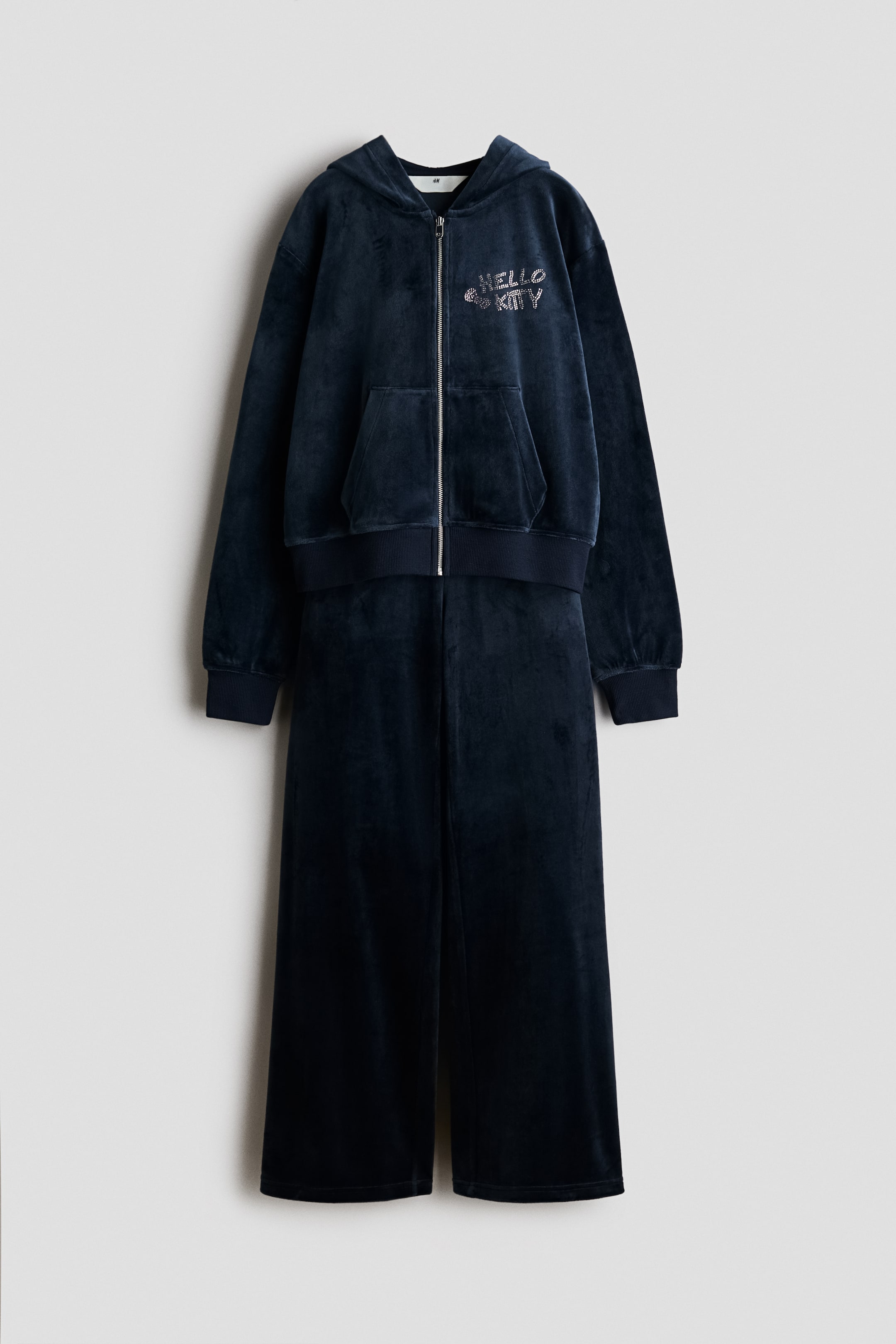 Größeres Bild ansehen: Eine Frontansicht eines passenden Sets aus dunkelmarineblauem Velours mit einer Kapuzenjacke mit Reißverschluss und einer Hose mit weitem Bein. Die Kapuzenjacke verfügt über einen silbernen Frontreißverschluss, Kängurutaschen, Rippbündchen an Ärmeln und Saum sowie ein Strass-„Hello Kitty“-Design auf der Brust, während die passende Hose gerade darunter hängt.