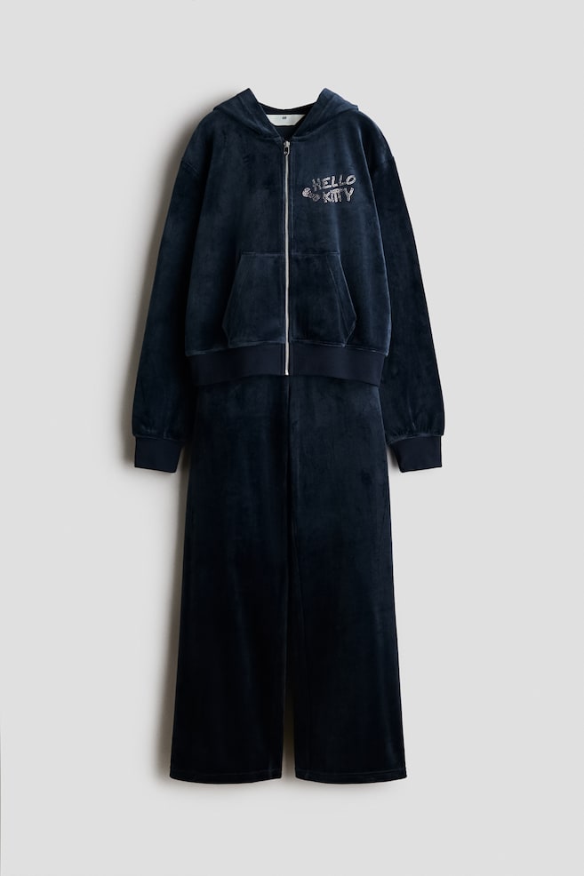 Vista frontal de un conjunto a juego de terciopelo azul marino oscuro que muestra una sudadera con capucha y cremallera y pantalones de pernera ancha. La sudadera presenta una cremallera frontal plateada, bolsillo canguro, puÃ±os y bajo de canalÃ©, y un diseÃ±o de âHello Kittyâ con pedrerÃ­a en el pecho, mientras que los pantalones a juego cuelgan rectos debajo. - 1
