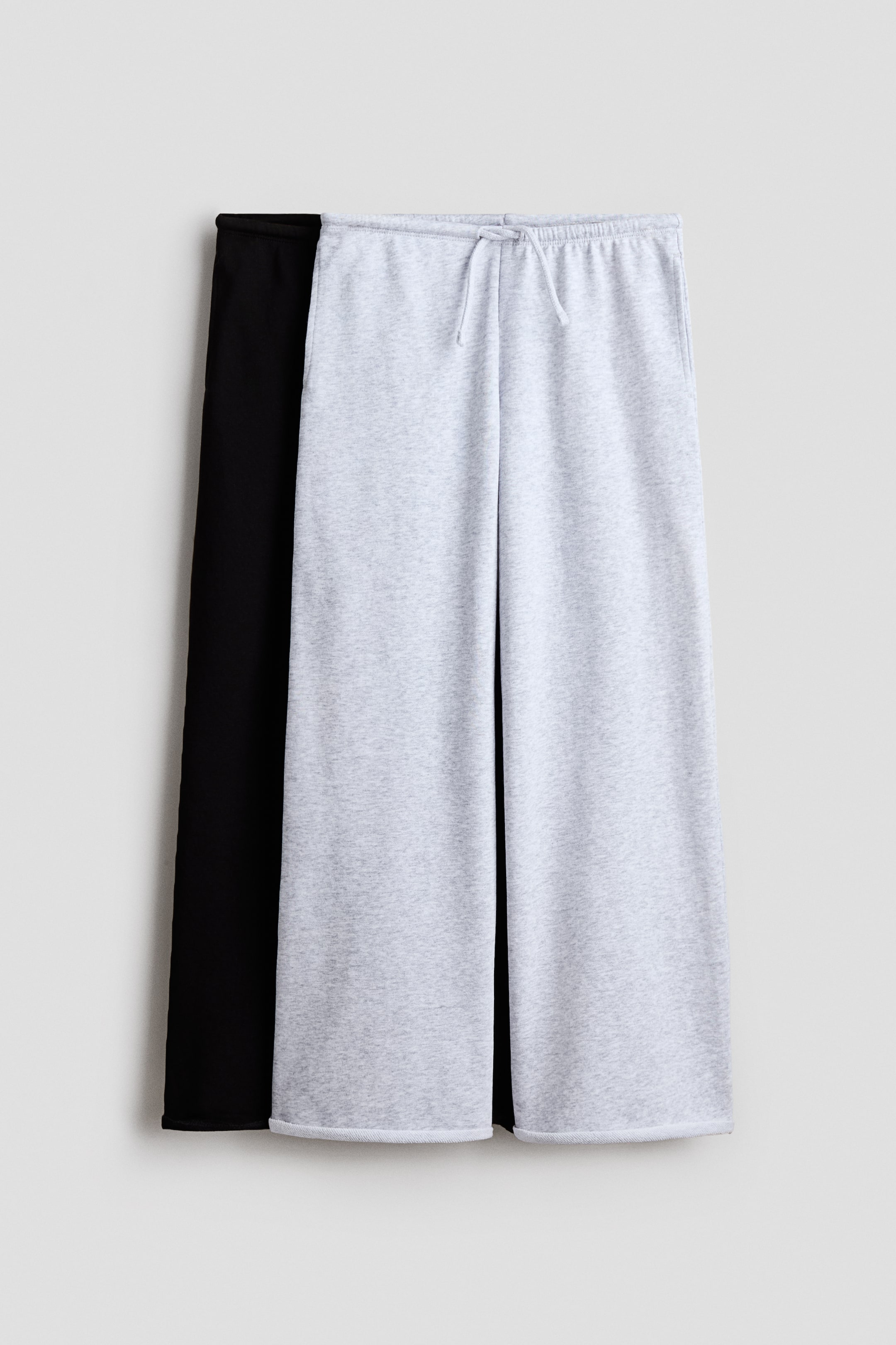 View larger image: A light gray marl pair of joggers, crafted from lightweight sweatshirt fabric, features an elasticated, drawstring waist, welt side pockets, wide legs, and roll-edge hems, with a black pair partially visible behind it.