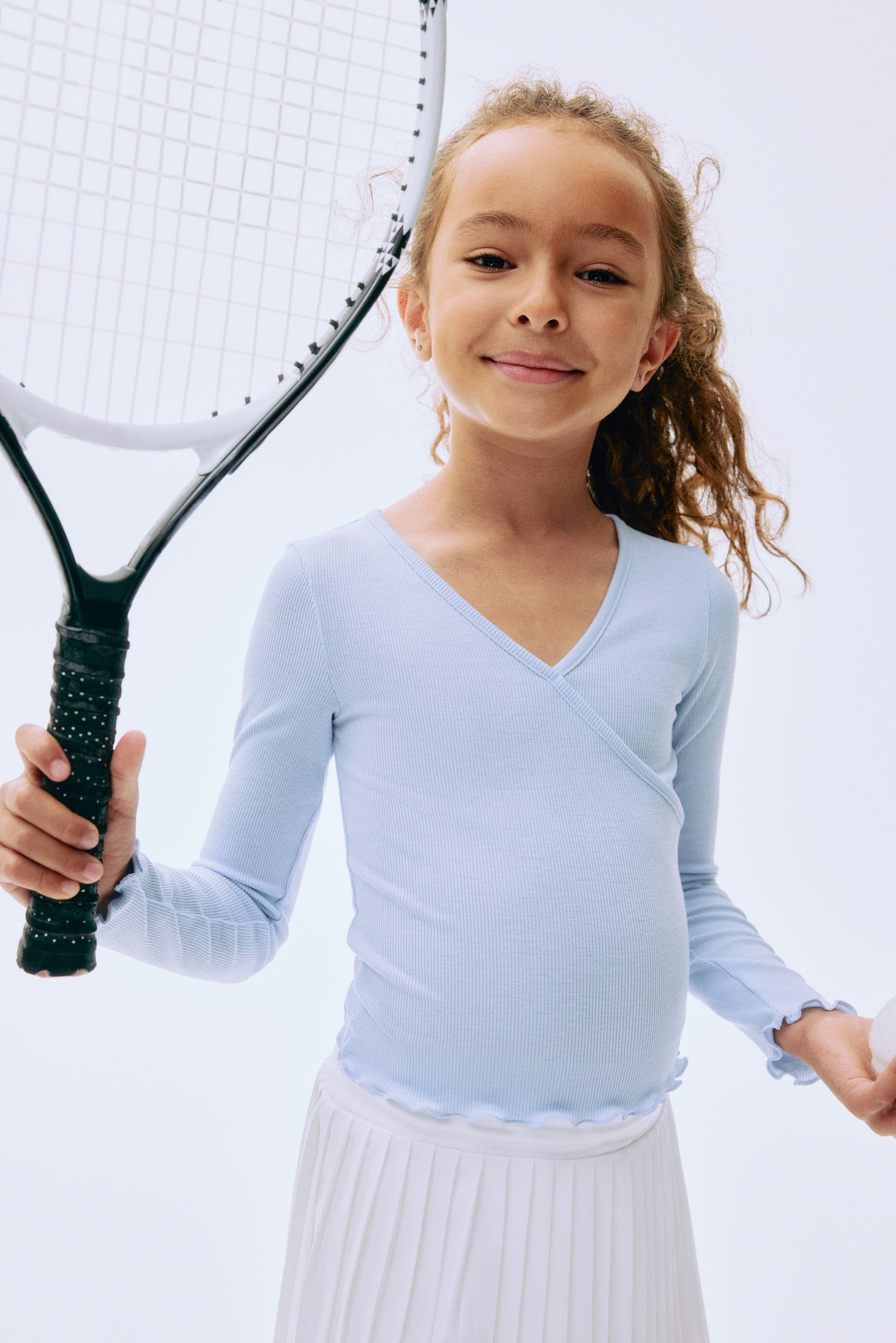 View larger image: Light blue ribbed long-sleeve top featuring a wrap-style V-neck with ruffle trim on the cuffs and hem. It layers over a white pleated skirt.