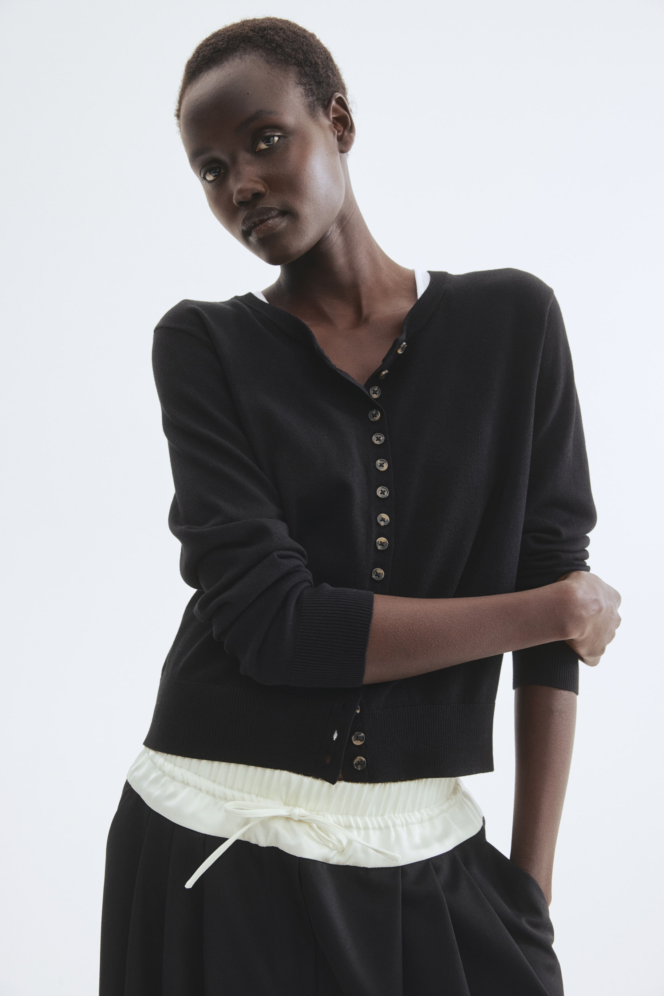 拡大画像を表示: 黒の細かいニットのカーディガンが、やや左に向けて前から展示されています。Vネックライン、軽い色の小さなボタンが並ぶ全体的なボタンプラケット、リブ付きのカフスの長袖、リブ付きの裾が特徴で、下に白い衣服が見えています。