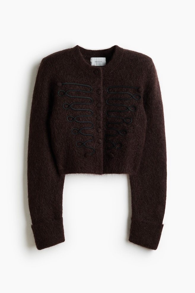 A dark brown, fuzzy-knit cropped cardigan is facing front, displaying a crew neck, a front button closure with covered buttons, and long sleeves with wide cuffs. Symmetrical, black cord embroidery in a military-inspired design is detailing both sides of the chest. - 1