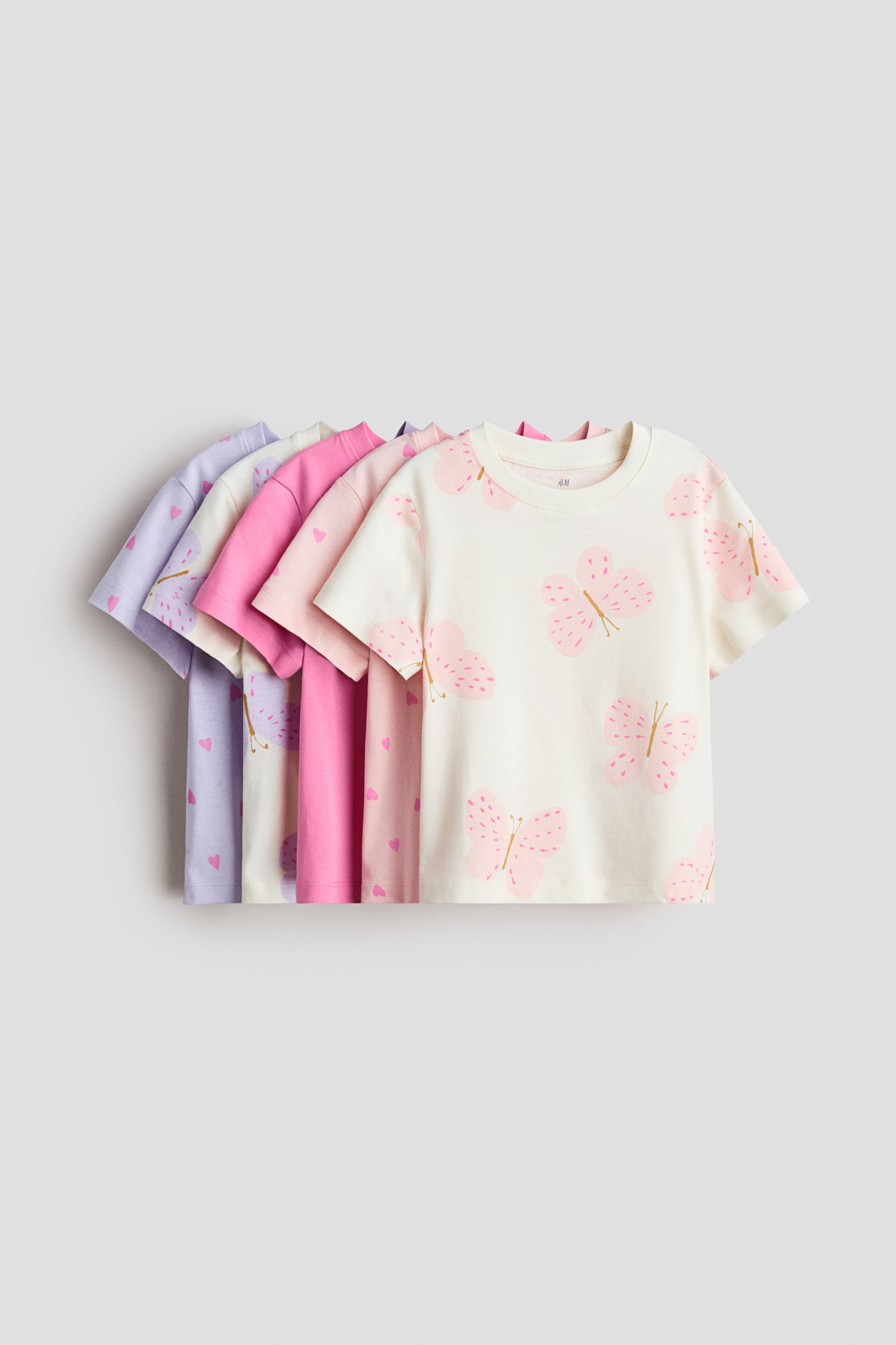 Visualizza immagine più grande: Cinque t-shirt in morbido jersey di cotone sono disposte in fila sovrapposta, con la t-shirt più avanti che presenta un colore bianco sporco con motivi di farfalle rosa, e le altre che mostrano un rosa chiaro con cuori, un rosa acceso a tinta unita e un viola chiaro con cuori, tutte caratterizzate da un bordo a costine intorno allo scollo.