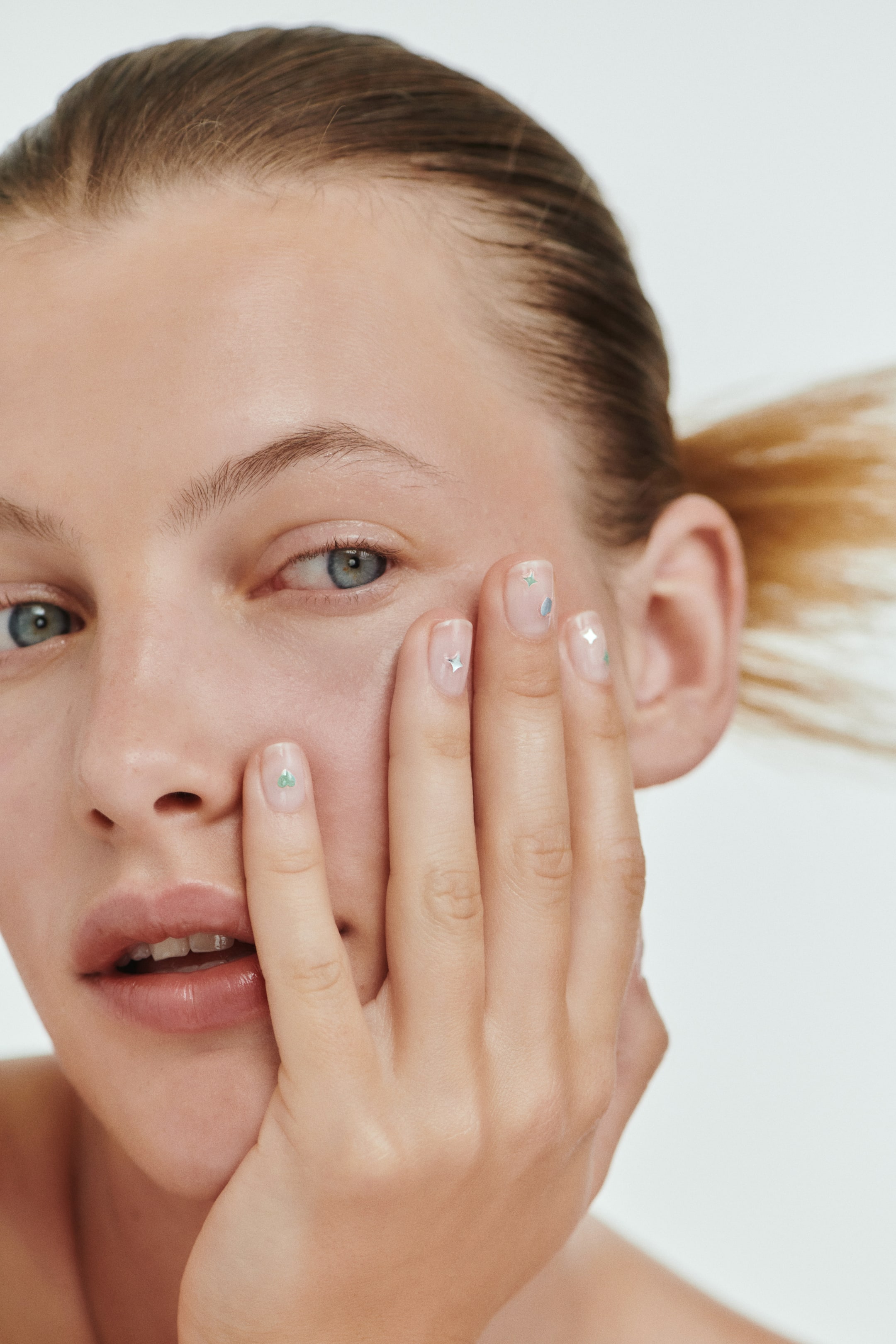 View larger image: 3D nail stickers, showcasing mint-coloured heart and star motifs alongside silver star designs, are adorned on the model's fingernails. The model is gently touching her face with her hand, highlighting the intricate manicure.