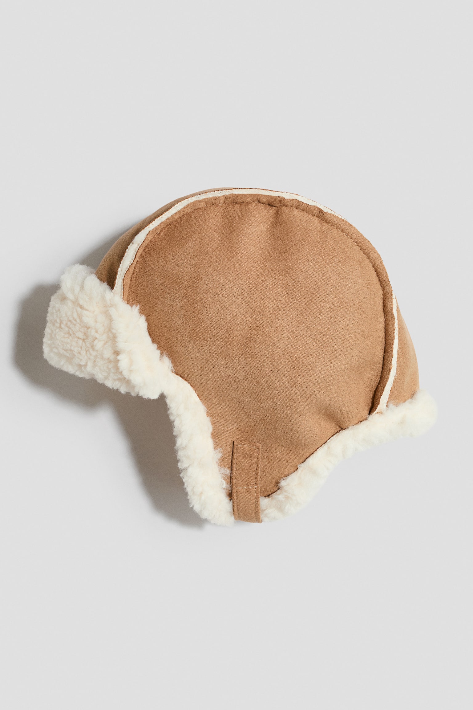Un gorro trapper de ante sintÃ©tico color tostado descansa de lado, mostrando un forro de pelo sintÃ©tico color crema que remata los bordes, las orejeras y el ala delantera doblada hacia arriba. El gorro presenta una construcciÃ³n de paneles curvos con ribeteado blanco visible y una pequeÃ±a presilla de tela color tostado en el lateral.