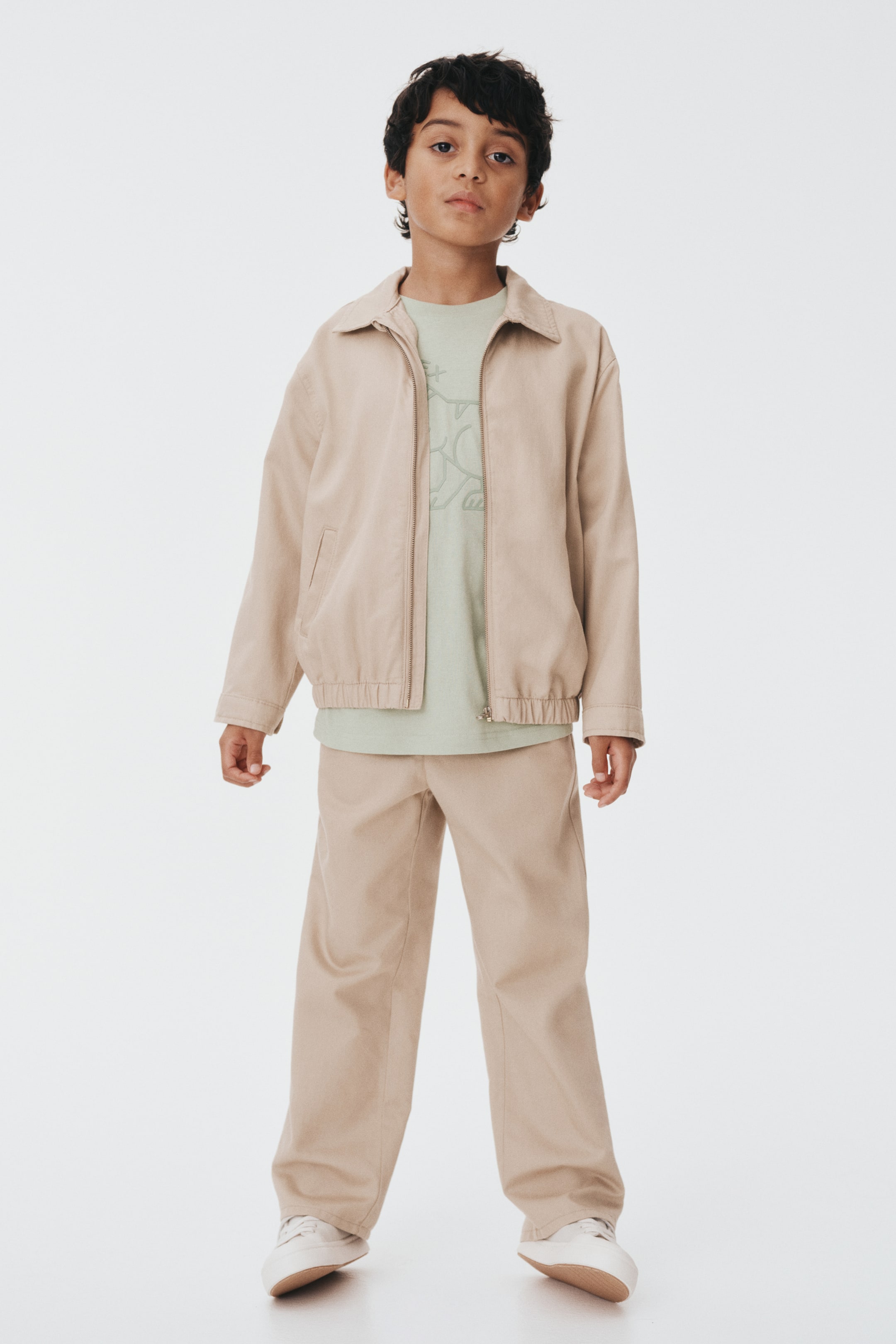 Büyük resmi görüntüle: Ensemble, öne doğru bakarken, yaygın bir yaka, tam fermuarlı bir ön ve düz manşetler ile yan ceplerle elastik bir etek ucu olan açık bej bir ceket sunuyor. Bu ceket, zarif bir grafik baskı sergileyen açık yeşil bir tişörtün üzerini örtüyor. Uyumlu açık bej geniş paçalı pantolonlar görünümü tamamlıyor.