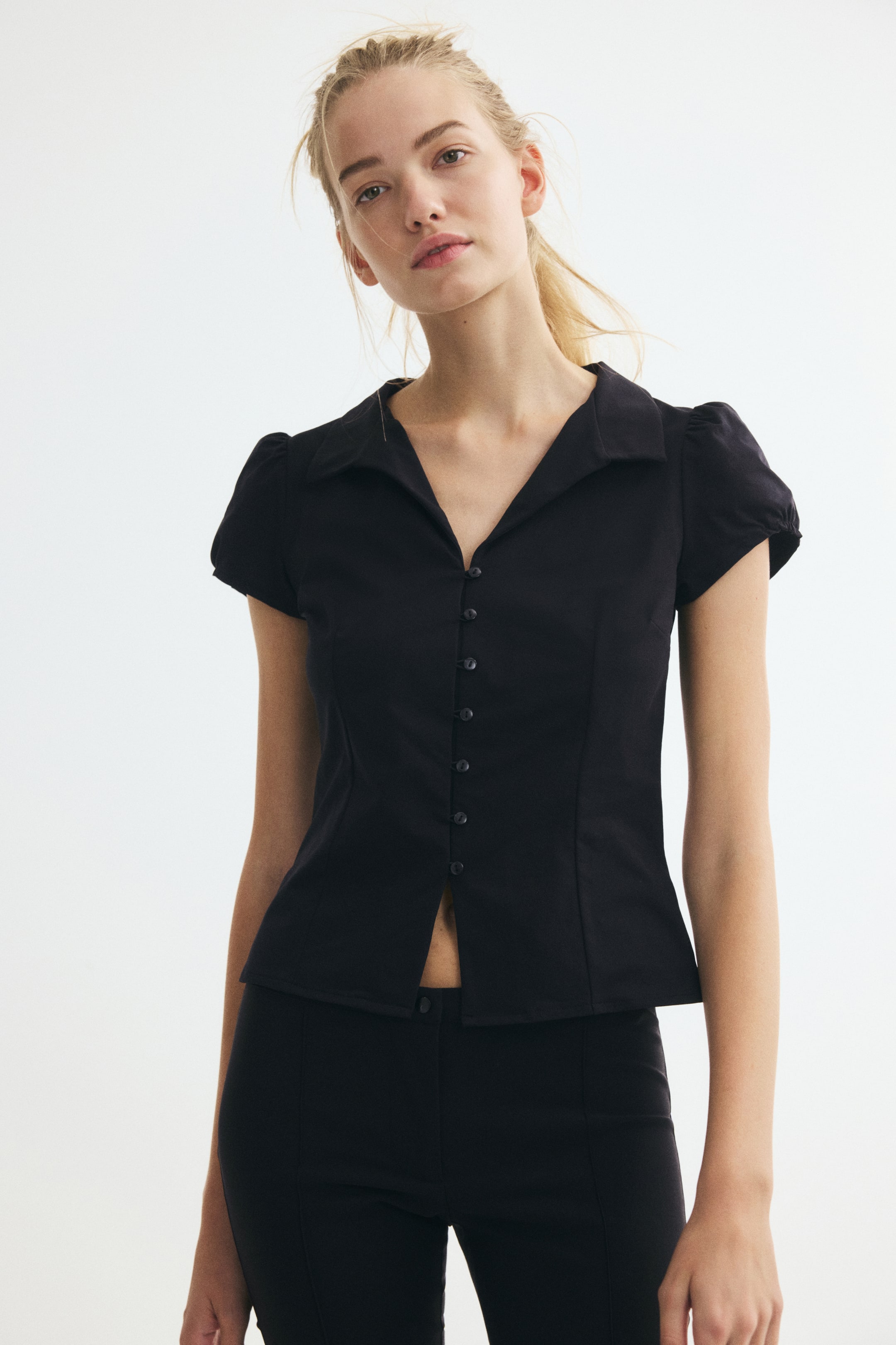 View larger image: The model is wearing a black, short-sleeved collared button-front top. The fitted top features puff sleeves, a V-neckline, and a button closure, with the lower buttons left open above matching black trousers.