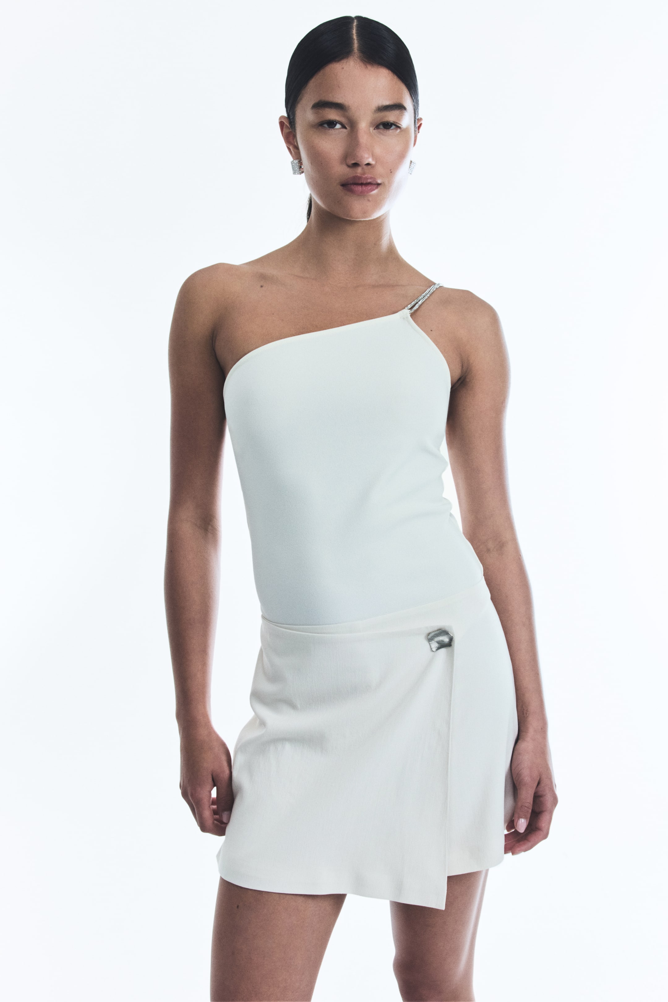 View larger image: A white one-shoulder top is featuring a rhinestone chain strap on the right shoulder. It is pairing with a white wrap mini-skirt, which is securing at the left hip with a silver-toned, geometric clasp.