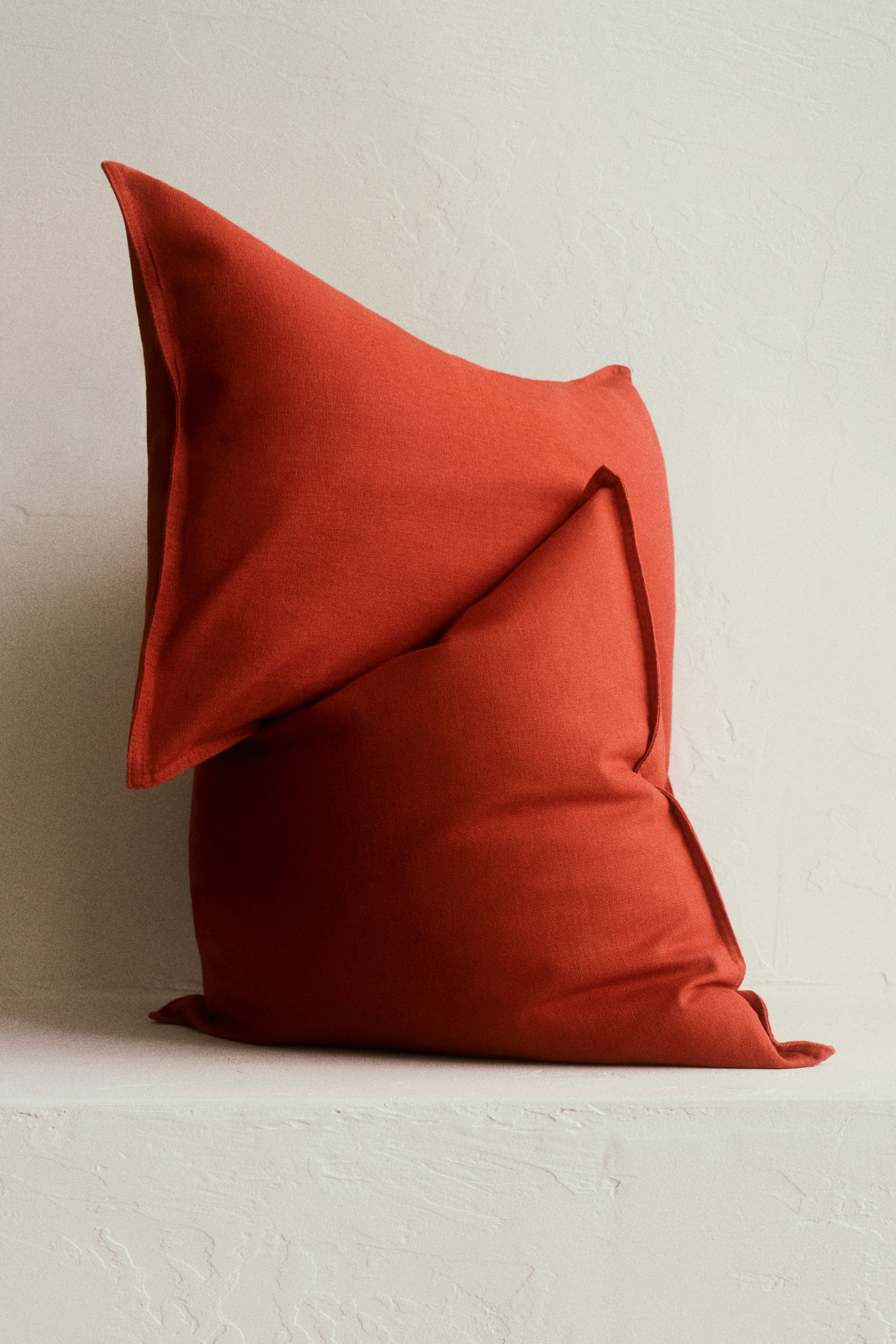 Voir une image plus grande: Deux coussins en tissu rouge sont disposés en une superposition, le coussin du dessus s'appuyant contre celui du dessous. Le coussin du dessus est incliné vers le haut, mettant en valeur ses coutures définies et ses bords nets.