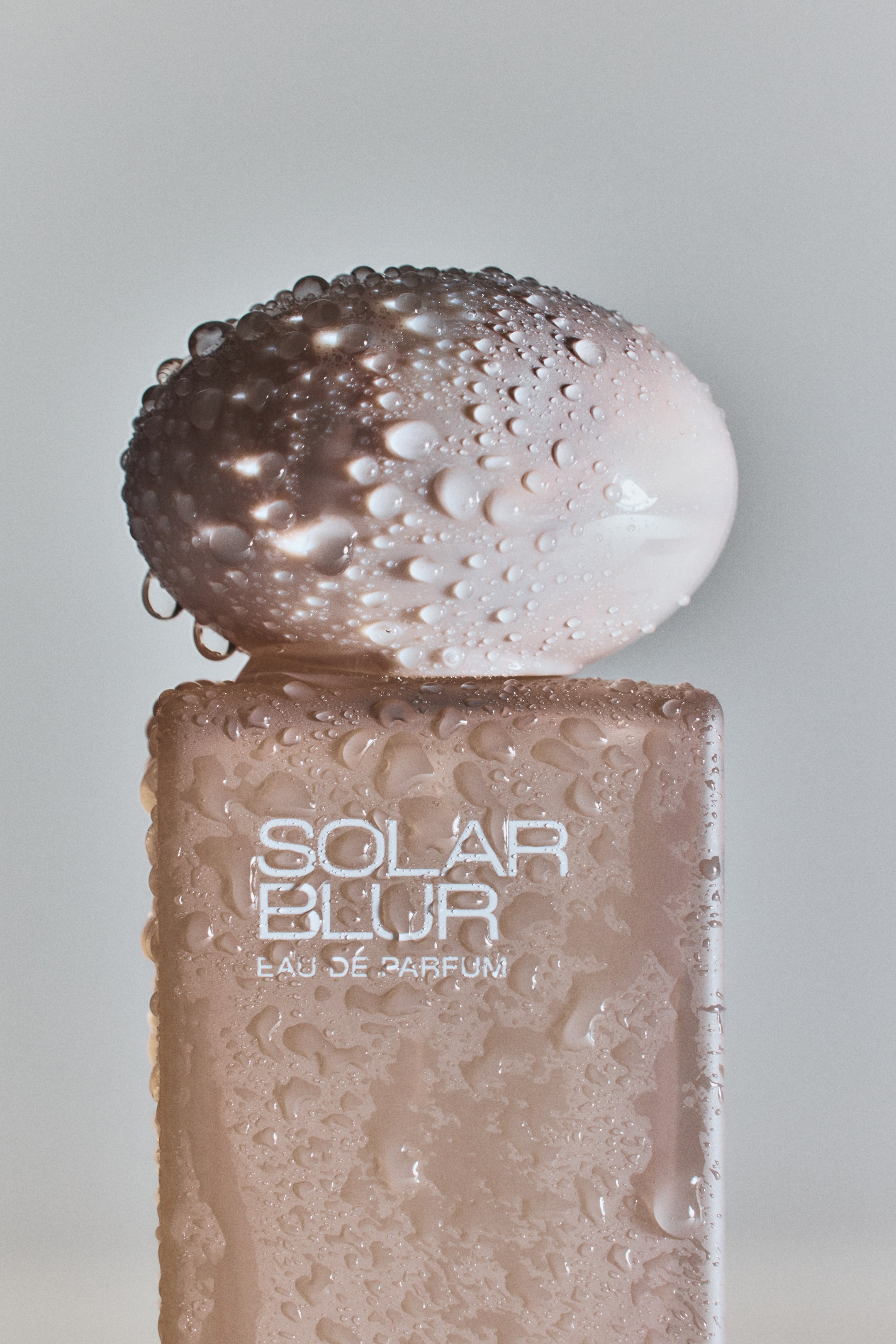 Grösseres Bild ansehen: Ein rechteckiges, hell pfirsichfarbenes Parfümfläschchen, das nach vorne zeigt, präsentiert den weißen Schriftzug "SOLAR BLUR EAU DE PARFUM" auf seiner Vorderseite. Ein abgerundeter, organisch geformter Verschluss ruht obenauf, und sowohl die Flasche als auch der Verschluss sind vollständig mit zahlreichen Wassertropfen bedeckt.