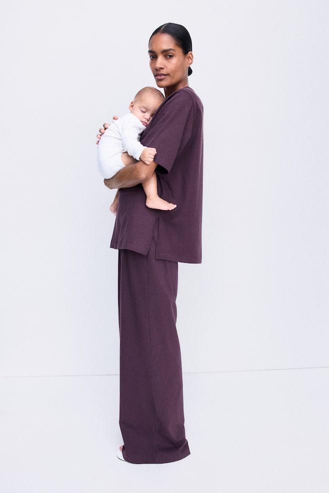La modelo lleva un conjunto de camiseta y pantalÃ³n en morado oscuro mientras sostiene a un bebÃ©. La camiseta de corte relajado presenta hombros caÃ­dos y bajo con aberturas laterales, combinada con un pantalÃ³n ancho a juego. - 1