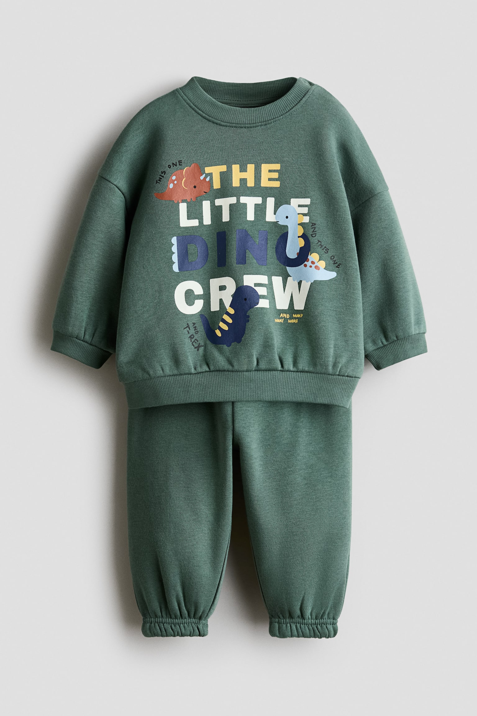 Má»t bá» Äá» ná» hai máº£nh mÃ u xanh lÃ¡ ÄÆ°á»£c chá»¥p tá»« phÃ­a trÆ°á»c, gá»m má»t Ã¡o sweatshirt tay dÃ i vÃ  quáº§n sweatpants Äá»ng bá». Ão sweatshirt cÃ³ cá» trÃ²n bo gÃ¢n, cá» tay bo gÃ¢n vÃ  gáº¥u Ã¡o bo gÃ¢n, vá»i há»a tiáº¿t in á» giá»¯a ghi "THE LITTLE DINO CREW" kÃ¨m hÃ¬nh minh há»a khá»§ng long, trong khi quáº§n sweatpants Äá»ng bá» cÃ³ pháº§n bo chun á» cá» chÃ¢n.