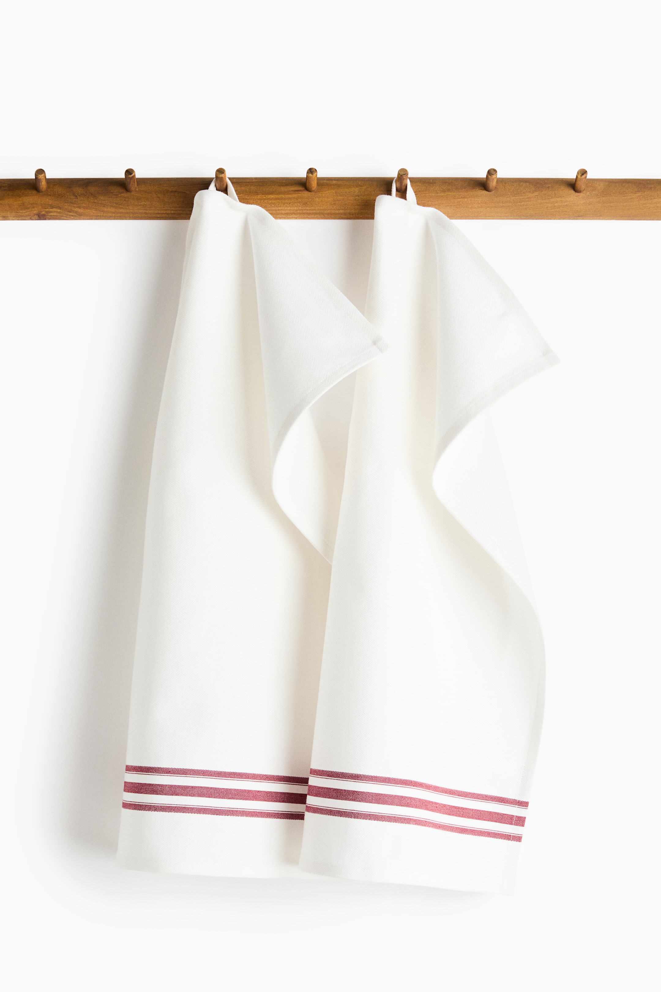 View larger image: Two off-white cotton tea towels in a herringbone weave are hanging vertically from a wooden peg rack by their loop hangers, displaying burgundy yarn-dyed horizontal stripes near the bottom hem.