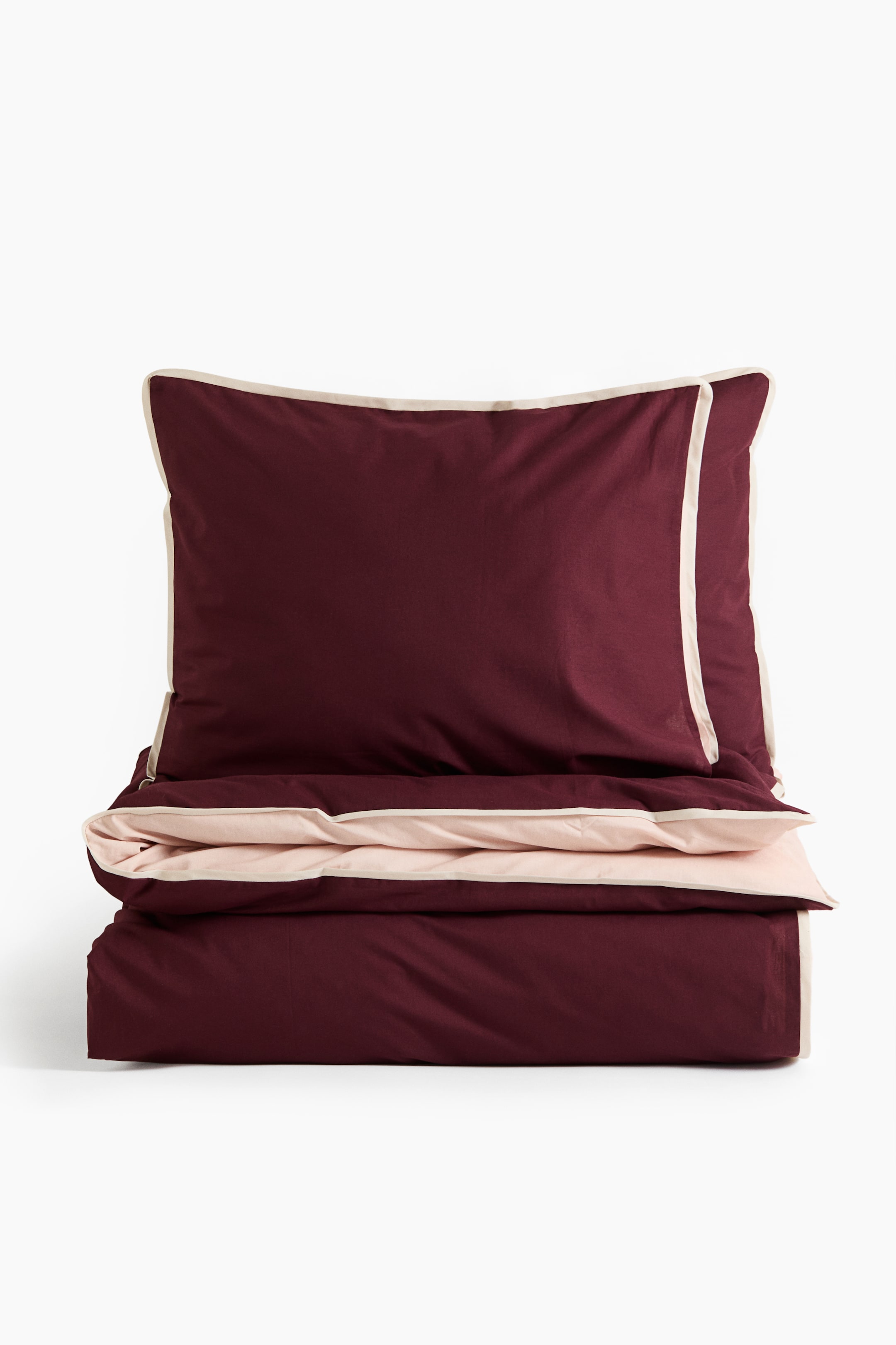 Agrandir l'image: Un ensemble de literie, présenté de face, comprend une taie d’oreiller bordeaux profond et une housse de couette repliée avec un envers rose pâle contrastant. Les deux pièces présentent un fin liseré passepoilé couleur crème le long de leurs bords.