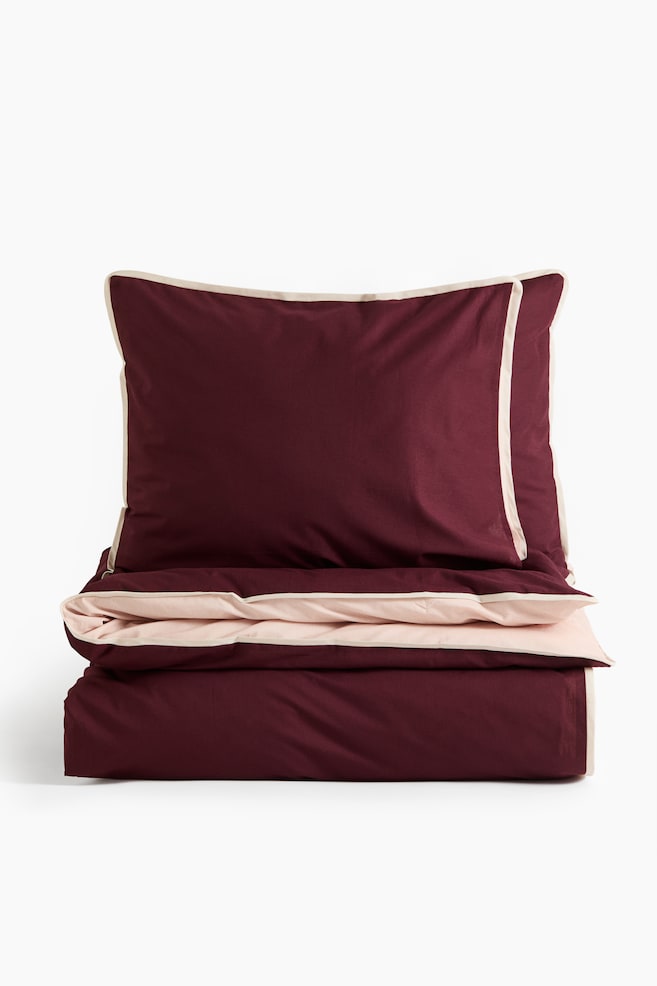 A bedding set, shown front-facing, features a deep burgundy pillowcase and a folded duvet cover with a contrasting light pink reverse side. Both pieces display a thin cream-coloured piped border along their edges. - 1