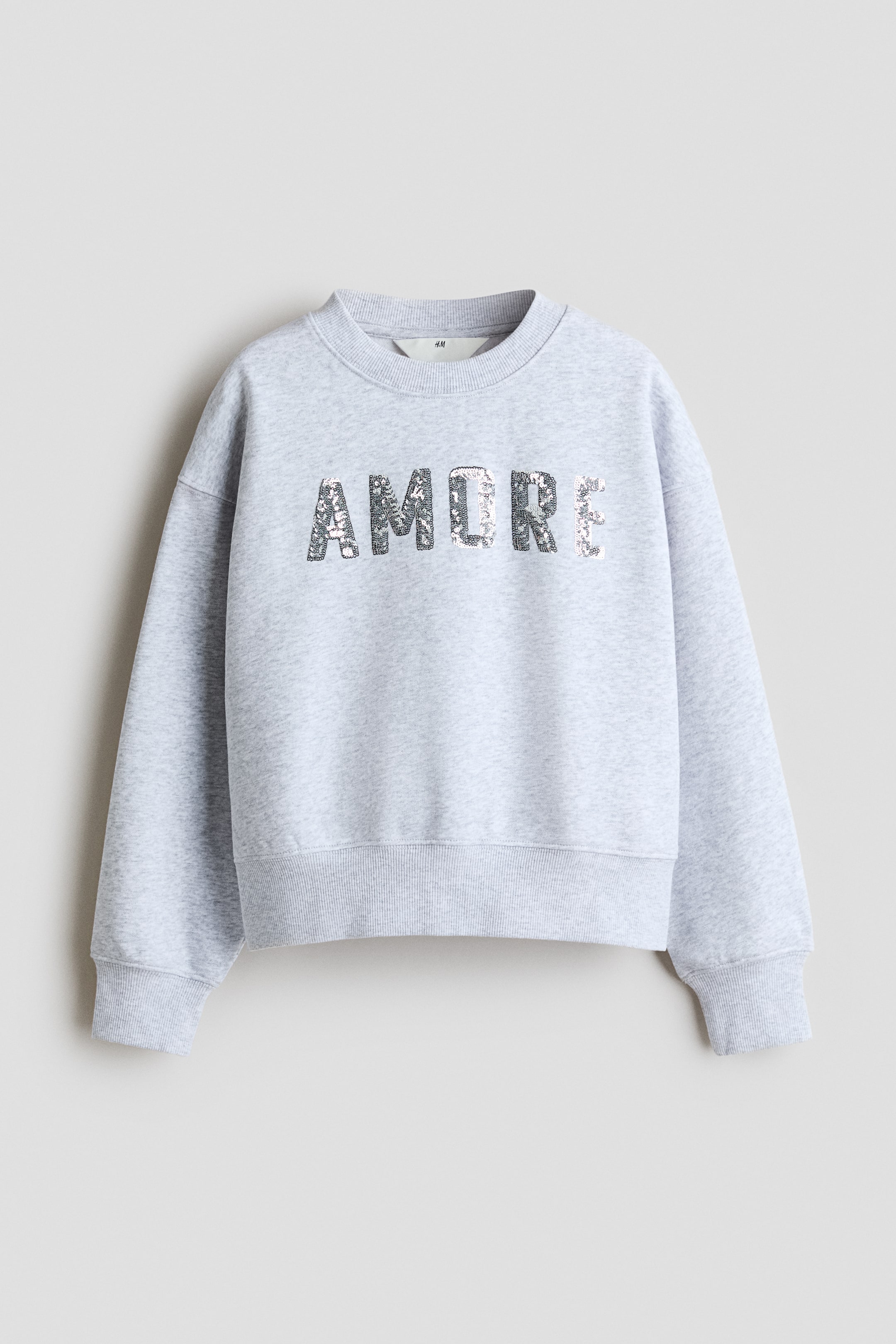 View larger image: A loose-fit top in grey fleece sweatshirt fabric is showcasing its front, adorned with the word "AMORE" in shimmering sequins. It features a round, rib-trimmed neckline, dropped shoulders, and ribbed cuffs and hem.