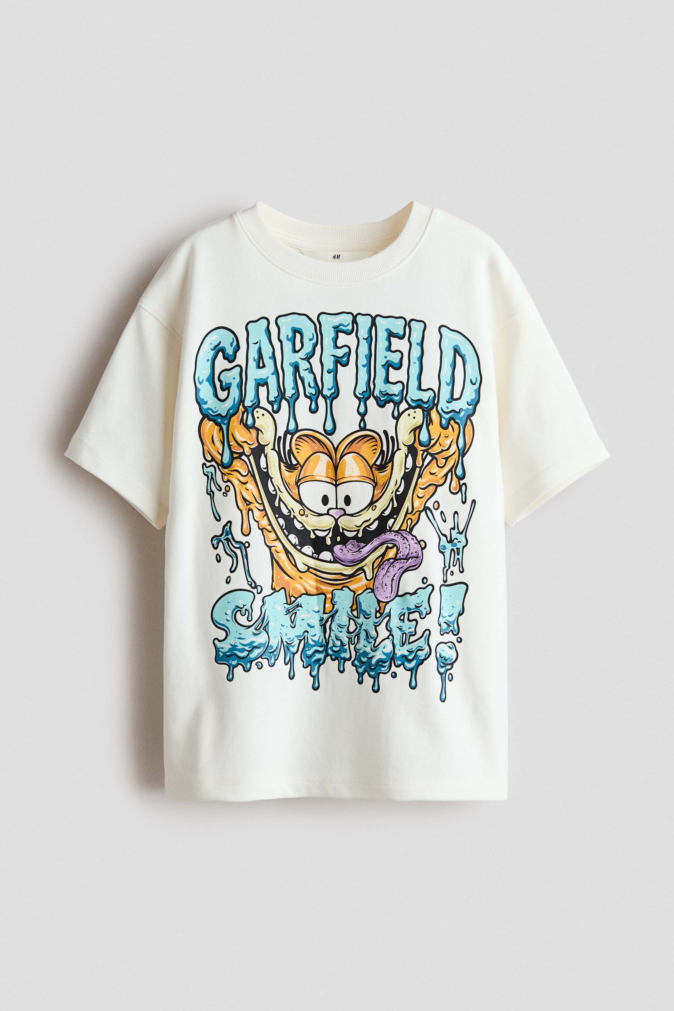 Visualizza l'immagine ingrandita: Una T-shirt a vestibilità ampia in tessuto felpato leggero e stampato presenta sul davanti una grande stampa grafica del viso sorridente di Garfield con testo blu e arancione che cola, recante le scritte "GARFIELD" e "SMILE!". È progettata con un girocollo rifinito a coste e spalle scese.