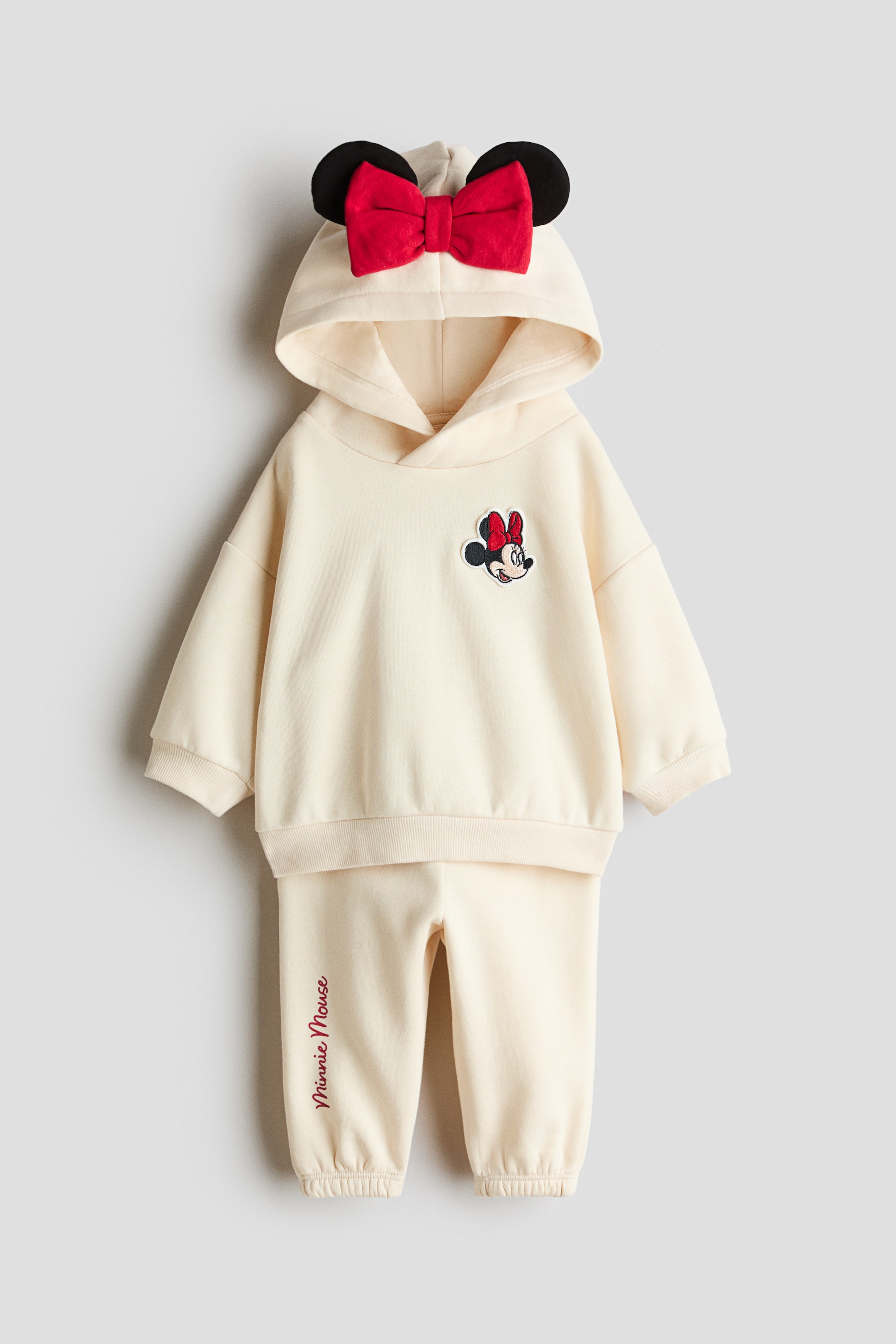 View larger image: A cream-colored hoodie and track pants set is shown from the front, featuring a wrap-front hood with attached black mouse ears and a large red bow. The hoodie has dropped shoulders, ribbed trims, and a Minnie Mouse patch on the chest, while the matching track pants feature elastic ankle cuffs and red embroidered text on the lower right leg.